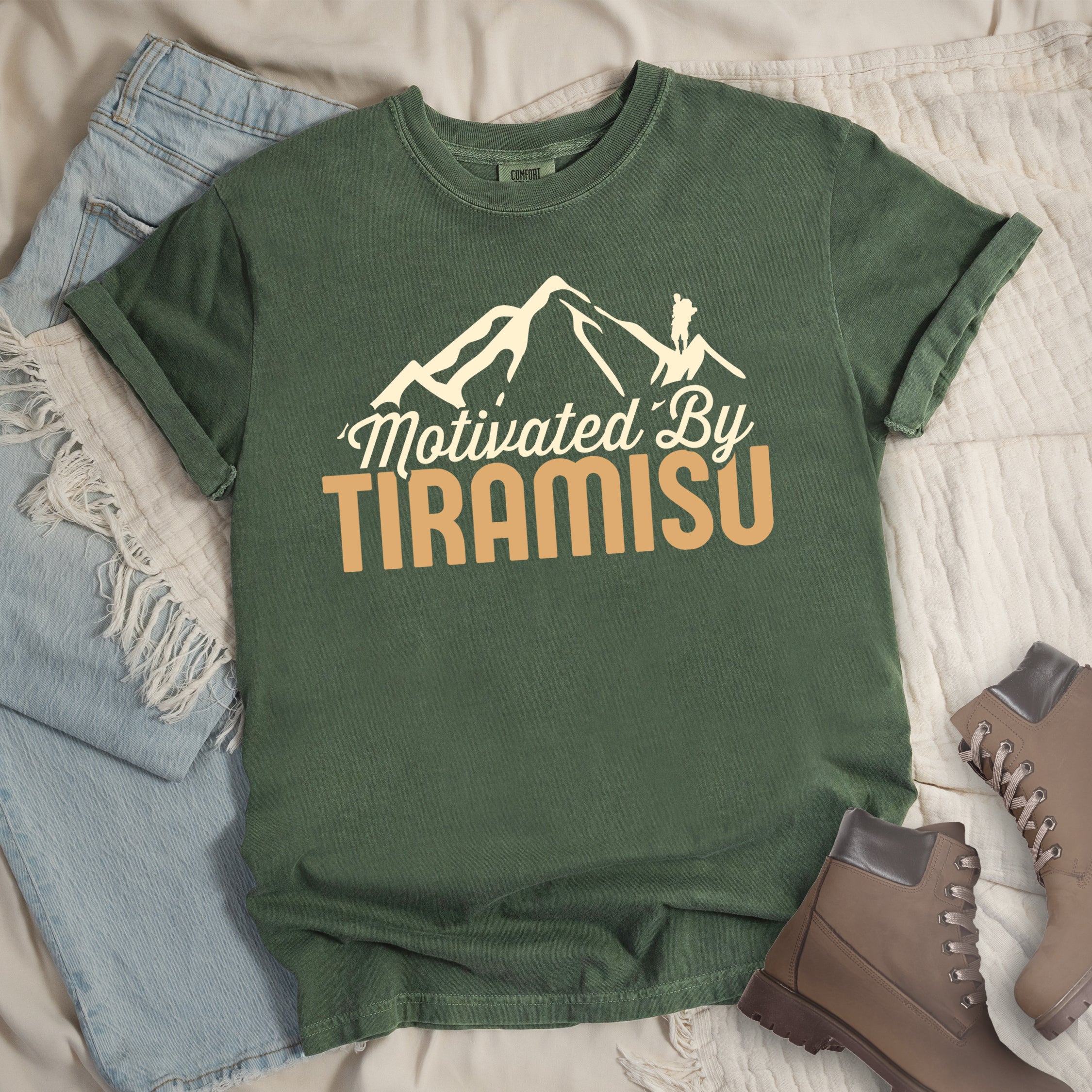 Moss (Dark Green) t-shirt with cream and gold text reading "Motivated By TIRAMISU". "Motivated By" appears in cream script underneath a cream mountain silhouette with a small hiker figure on the peak, and "TIRAMISU" in large gold letters at the bottom. The shirt features a classic crew neck and casual rolled sleeves, styled alongside light wash denim and brown hiking boots. The design humorously connects the Italian coffee-flavored dessert as motivation for hiking and outdoor activities.