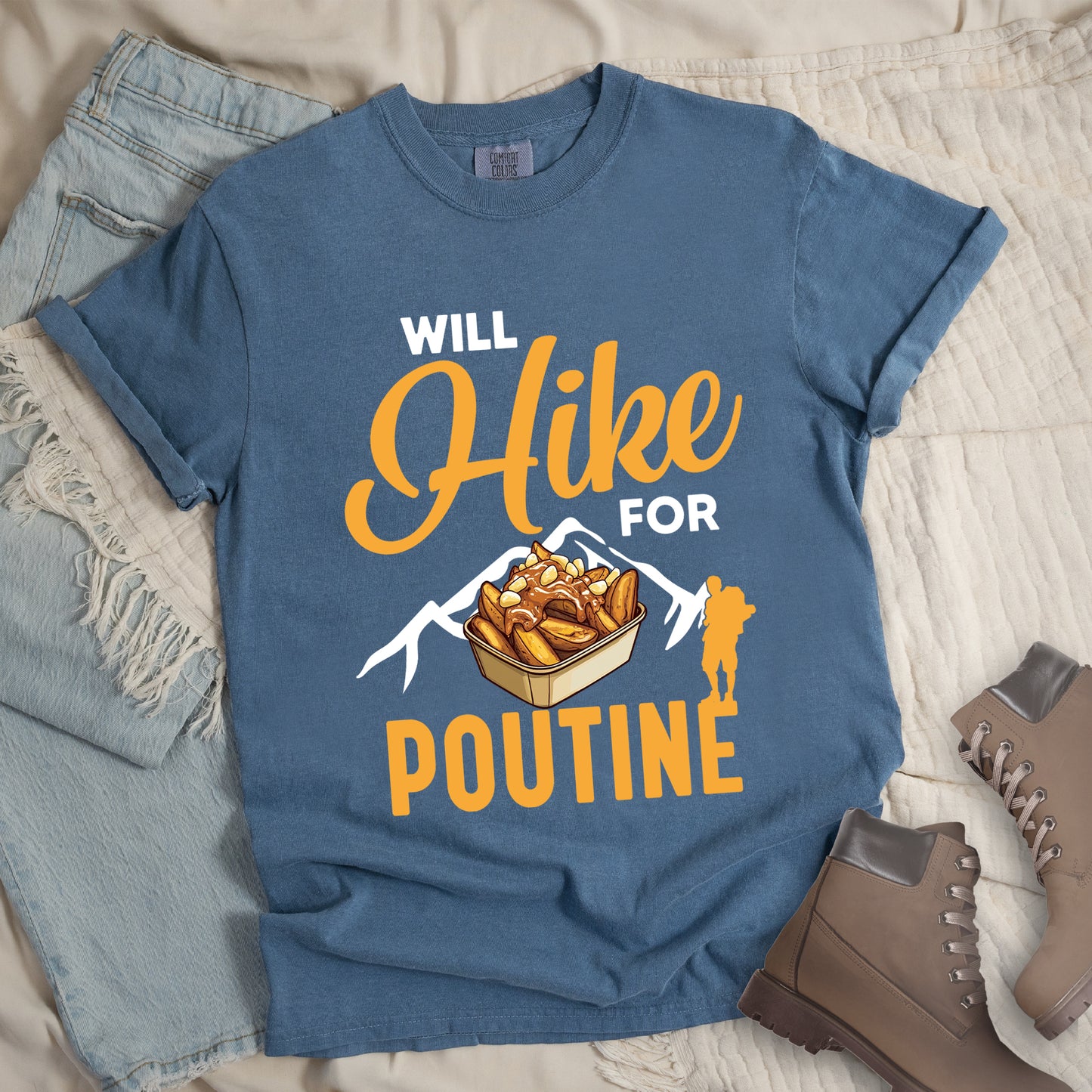Blue Jean (Denim Blue) t-shirt with orange and white text reading "WILL Hike FOR POUTINE". "WILL" appears in white at the top, "Hike" in large orange script in the middle, "FOR" in white, and "POUTINE" in large orange letters at the bottom. Features a detailed illustration of a container of poutine (french fries with cheese curds and gravy) in the center, white mountain silhouettes behind it, and an orange hiker silhouette on the right.