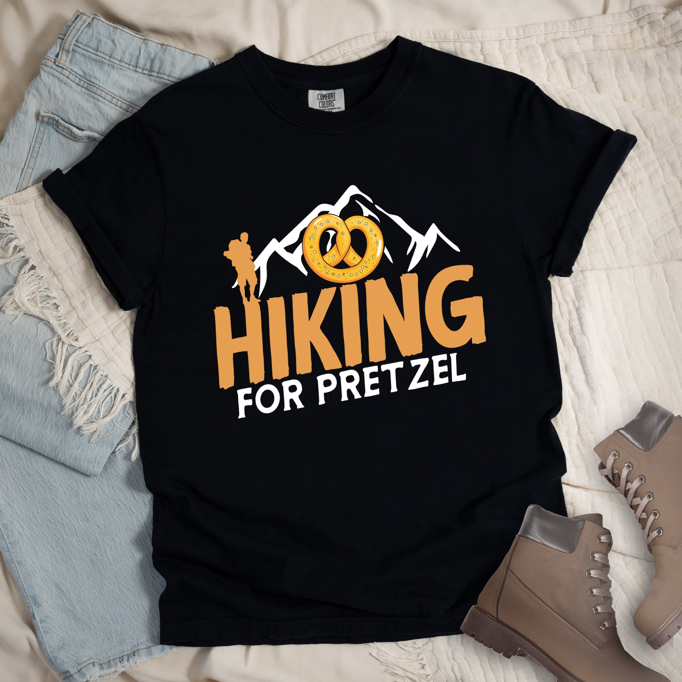 Black t-shirt with orange and white text reading "HIKING FOR PRETZEL". "HIKING" appears in large orange letters in the middle, and "FOR PRETZEL" in white below it. Features white mountain silhouettes in the background, an orange hiker silhouette on the left side, and an illustration of a golden pretzel with salt above the word "HIKING". The design connects hiking activity with pretzel snacks as a reward.