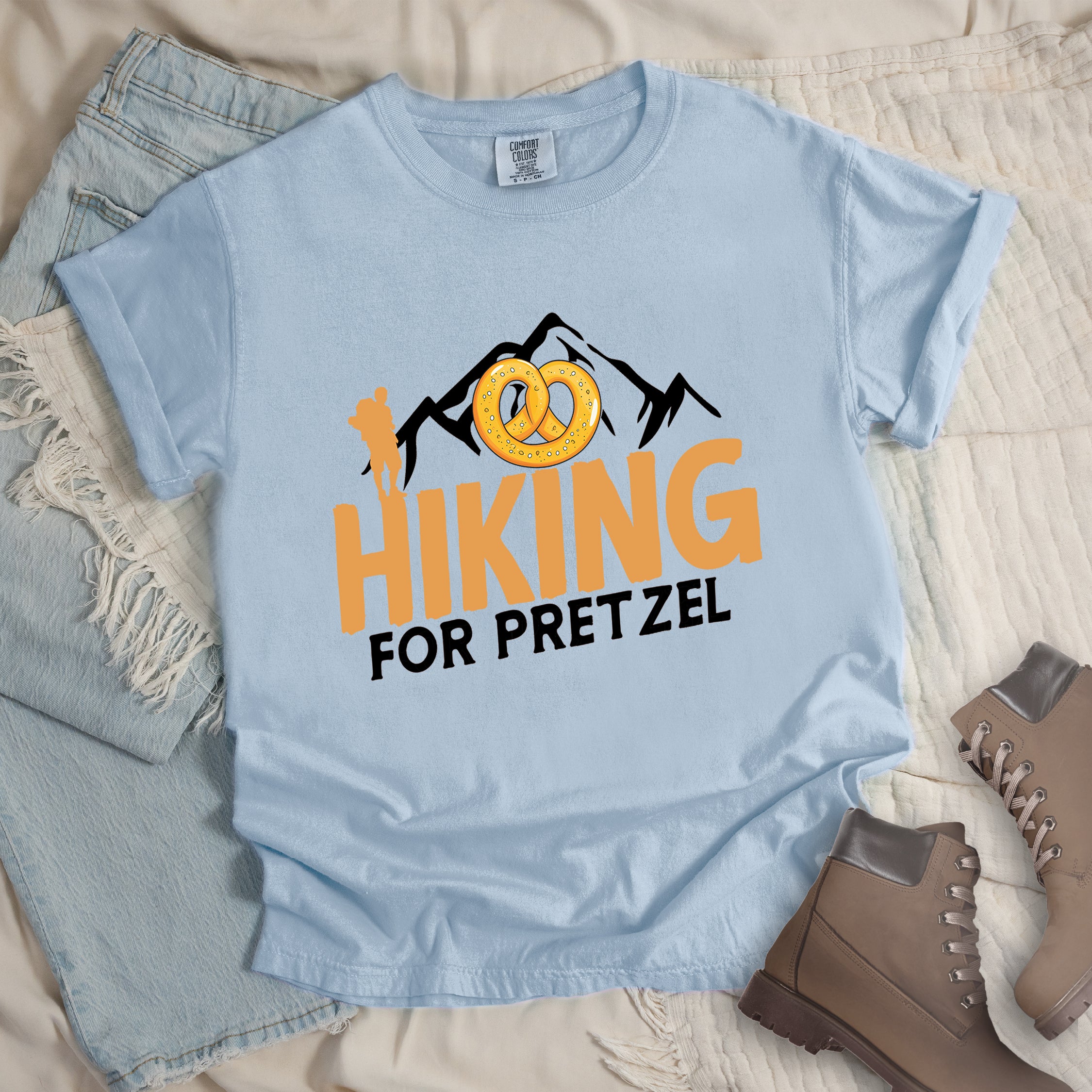 Chambray (Light Blue) t-shirt with orange and black text reading "HIKING FOR PRETZEL". "HIKING" appears in large orange letters in the middle, and "FOR PRETZEL" in black below it. Features black mountain silhouettes in the background, an orange hiker silhouette on the left side, and an illustration of a golden pretzel with salt above the word "HIKING". The design connects hiking activity with pretzel snacks as a reward.