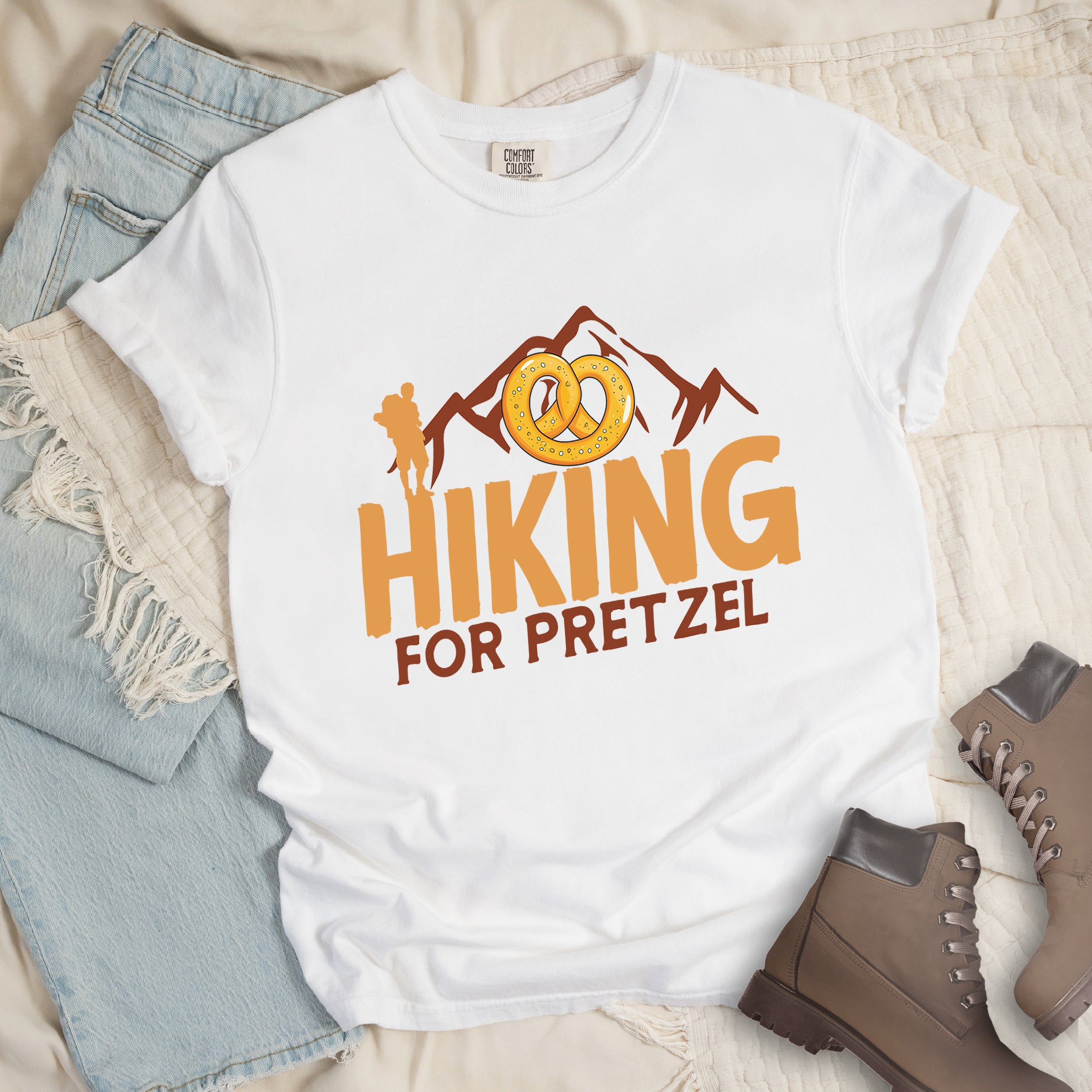 White t-shirt with orange and brown text reading "HIKING FOR PRETZEL". "HIKING" appears in large orange letters in the middle, and "FOR PRETZEL" in brown below it. Features brown mountain silhouettes in the background, an orange hiker silhouette on the left side, and an illustration of a golden pretzel with salt above the word "HIKING". The design connects hiking activity with pretzel snacks as a reward.