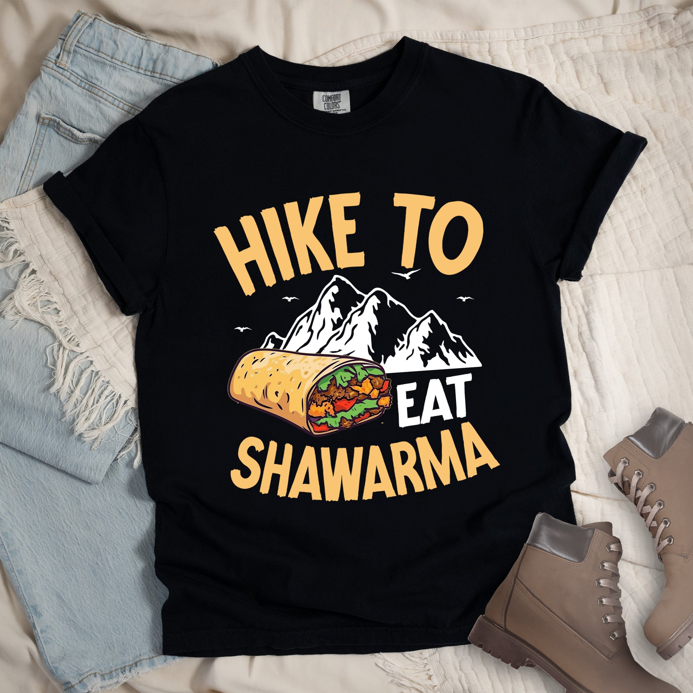 Black t-shirt with golden yellow and white text reading "HIKE TO EAT SHAWARMA". "HIKE TO" appears in golden yellow letters at the top, "EAT" in white in the middle, and "SHAWARMA" in golden yellow at the bottom. Features a detailed illustration of a shawarma wrap with visible meat and vegetables, white mountain silhouettes with small bird silhouettes in the background. Features a classic crew neck and casual rolled sleeves, styled alongside light wash denim and brown hiking boots.