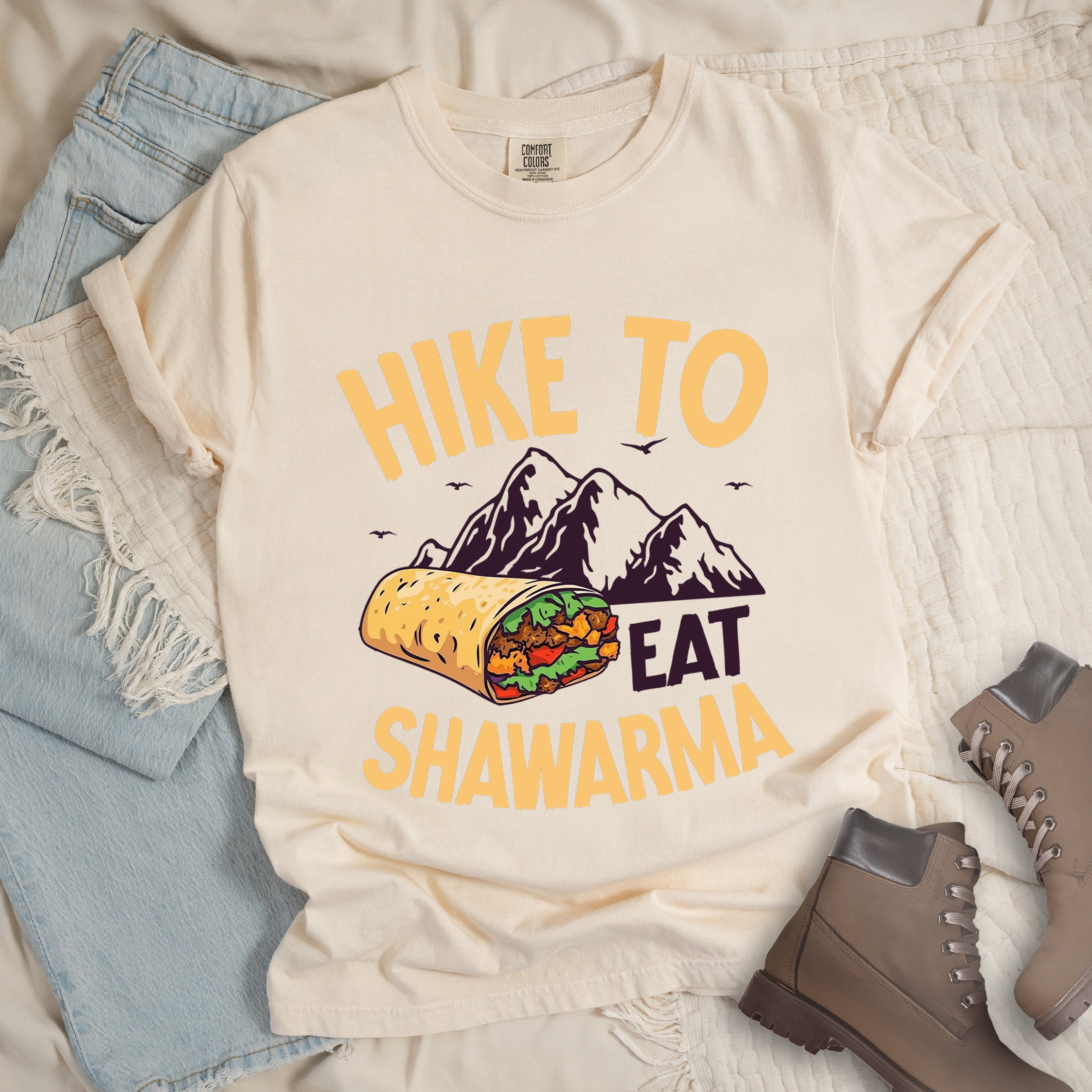 Ivory (Cream White) t-shirt with golden yellow and deep purple text reading "HIKE TO EAT SHAWARMA". "HIKE TO" appears in golden yellow letters at the top, "EAT" in deep purple in the middle, and "SHAWARMA" in golden yellow at the bottom. Features a detailed illustration of a shawarma wrap with visible meat and vegetables, deep purple mountain silhouettes with small bird silhouettes in the background.