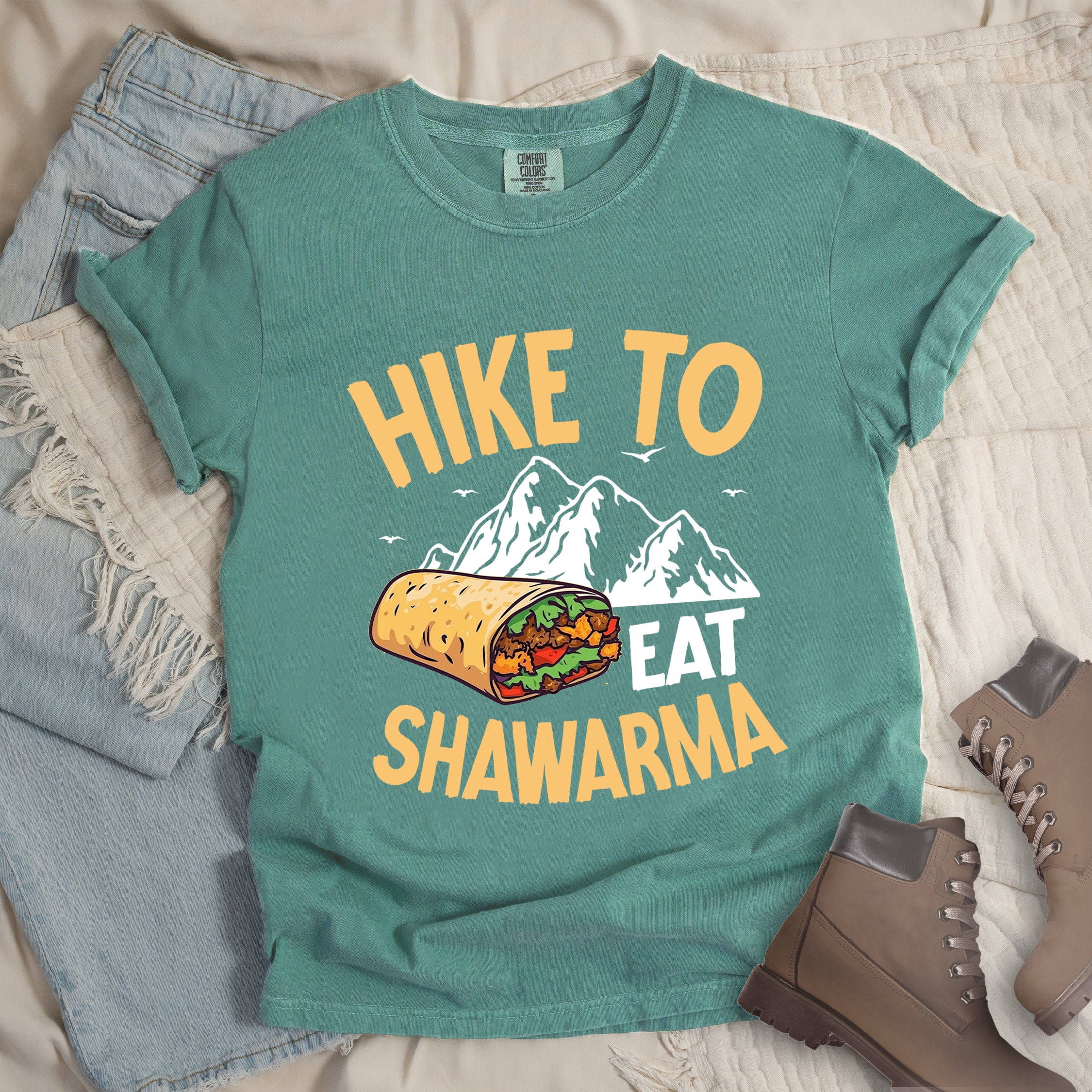 Light Green t-shirt with golden yellow and white text reading "HIKE TO EAT SHAWARMA". "HIKE TO" appears in golden yellow letters at the top, "EAT" in white in the middle, and "SHAWARMA" in golden yellow at the bottom. Features a detailed illustration of a shawarma wrap with visible meat and vegetables, white mountain silhouettes with small bird silhouettes in the background. Features a classic crew neck and casual rolled sleeves, styled alongside light wash denim and brown hiking boots.