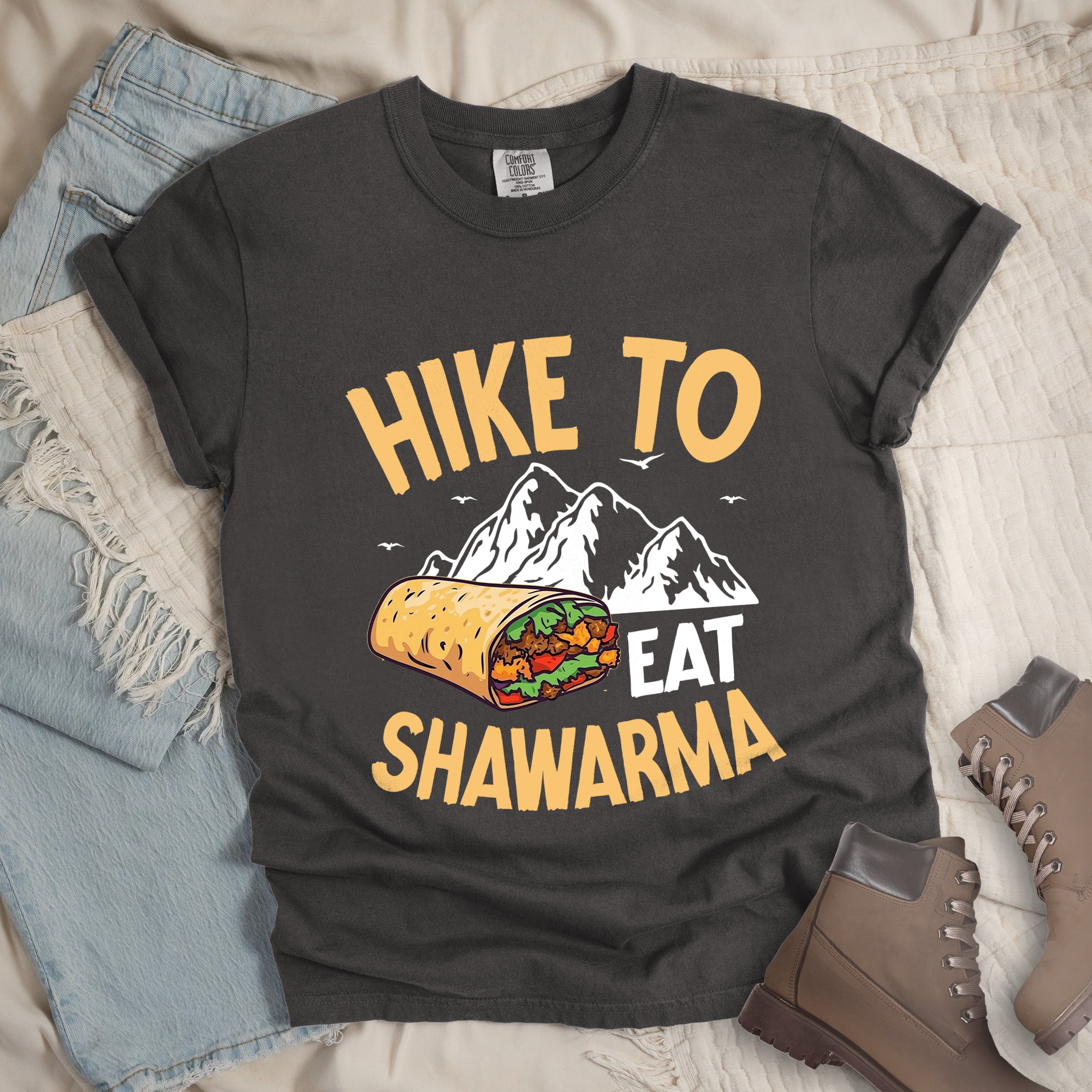 Pepper (Dark Gray) t-shirt with golden yellow and white text reading "HIKE TO EAT SHAWARMA". "HIKE TO" appears in golden yellow letters at the top, "EAT" in white in the middle, and "SHAWARMA" in golden yellow at the bottom. Features a detailed illustration of a shawarma wrap with visible meat and vegetables, white mountain silhouettes with small bird silhouettes in the background. Features a classic crew neck and casual rolled sleeves, styled alongside light wash denim and brown hiking boots.