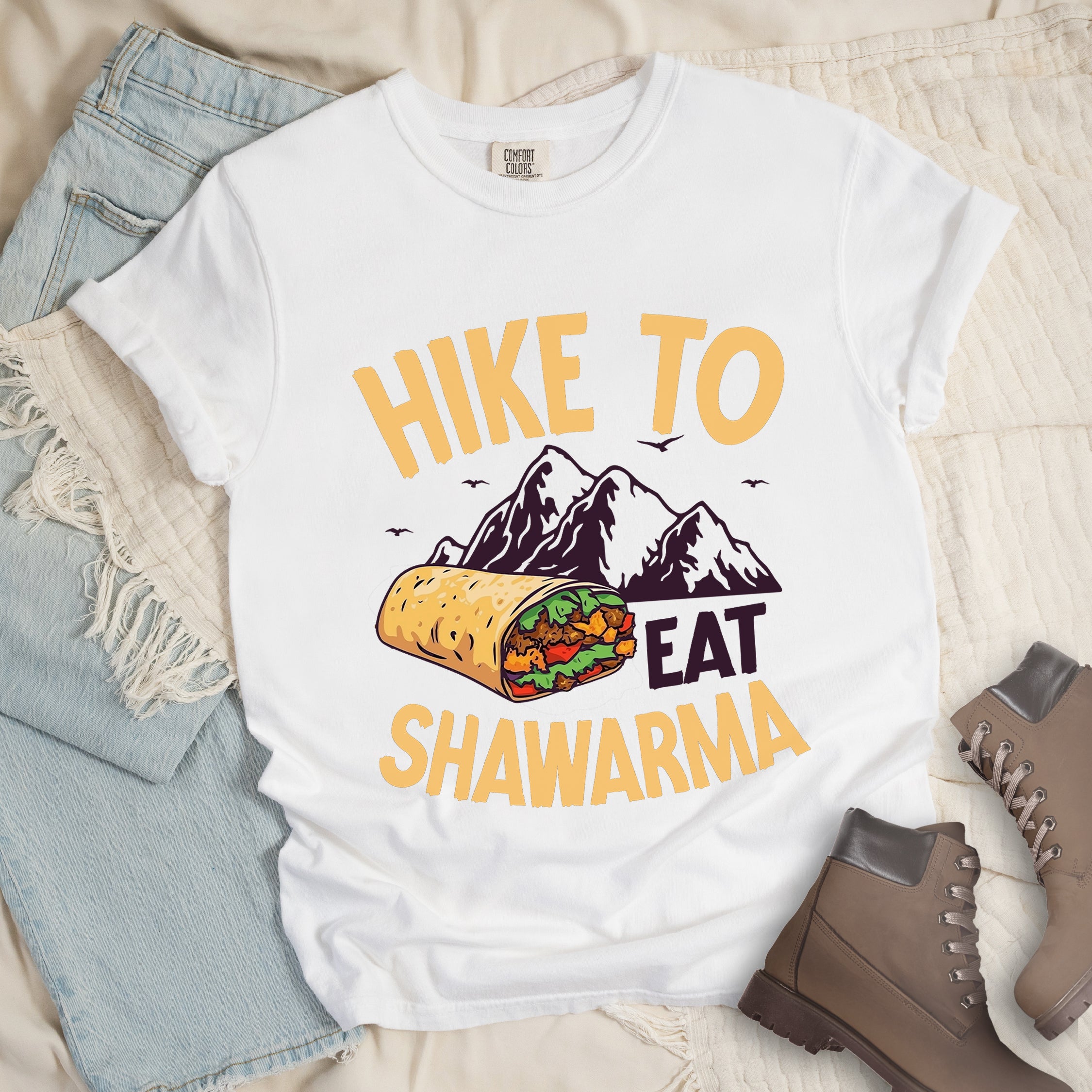 White t-shirt with golden yellow and deep purple text reading "HIKE TO EAT SHAWARMA". "HIKE TO" appears in golden yellow letters at the top, "EAT" in deep purple in the middle, and "SHAWARMA" in golden yellow at the bottom. Features a detailed illustration of a shawarma wrap with visible meat and vegetables, deep purple mountain silhouettes with small bird silhouettes in the background. Features a classic crew neck and casual rolled sleeves, styled alongside light wash denim and brown hiking boots.