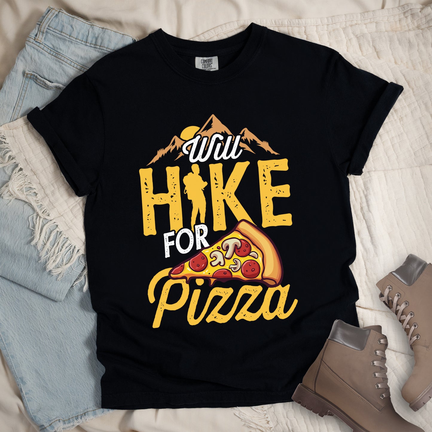Black t-shirt with "Will HIKE FOR pizza" design. Features "Will" in white script above mountain graphics with a yellow sun. "HIKE" appears in large yellow distressed letters with a hiker silhouette replacing the letter "I". "FOR" in white text sits above "pizza" in yellow script. An illustrated pizza slice with pepperoni and mushrooms appears between "FOR" and "pizza". The shirt features a classic crew neck and casual rolled sleeves, styled alongside light wash denim and brown hiking boots.