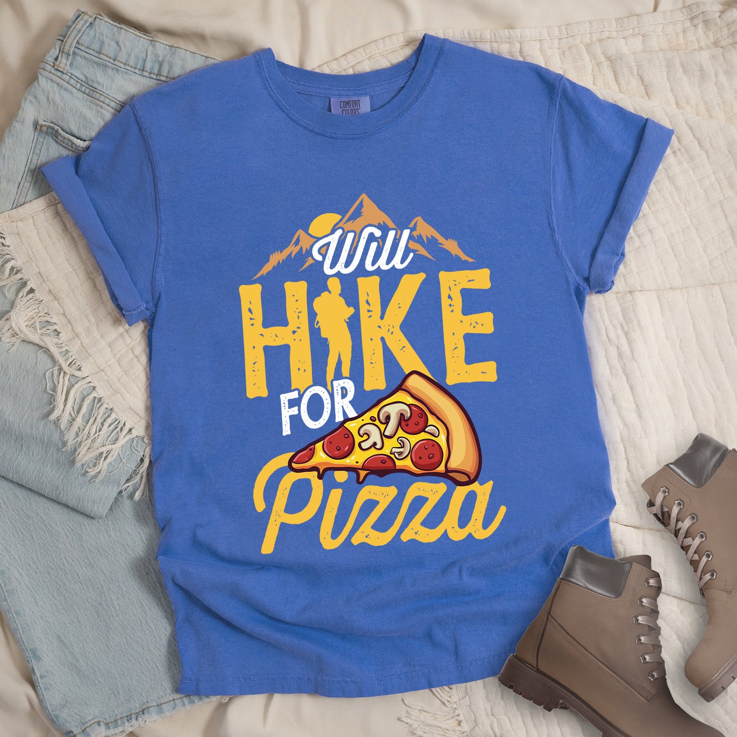 Flo Jean (Blue) t-shirt with "Will HIKE FOR pizza" design. Features "Will" in white script above mountain graphics with a yellow sun. "HIKE" appears in large yellow distressed letters with a hiker silhouette replacing the letter "I". "FOR" in white text sits above "pizza" in yellow script. An illustrated pizza slice with pepperoni and mushrooms appears between "FOR" and "pizza". The shirt features a classic crew neck and casual rolled sleeves, styled alongside light wash denim and brown hiking boots.