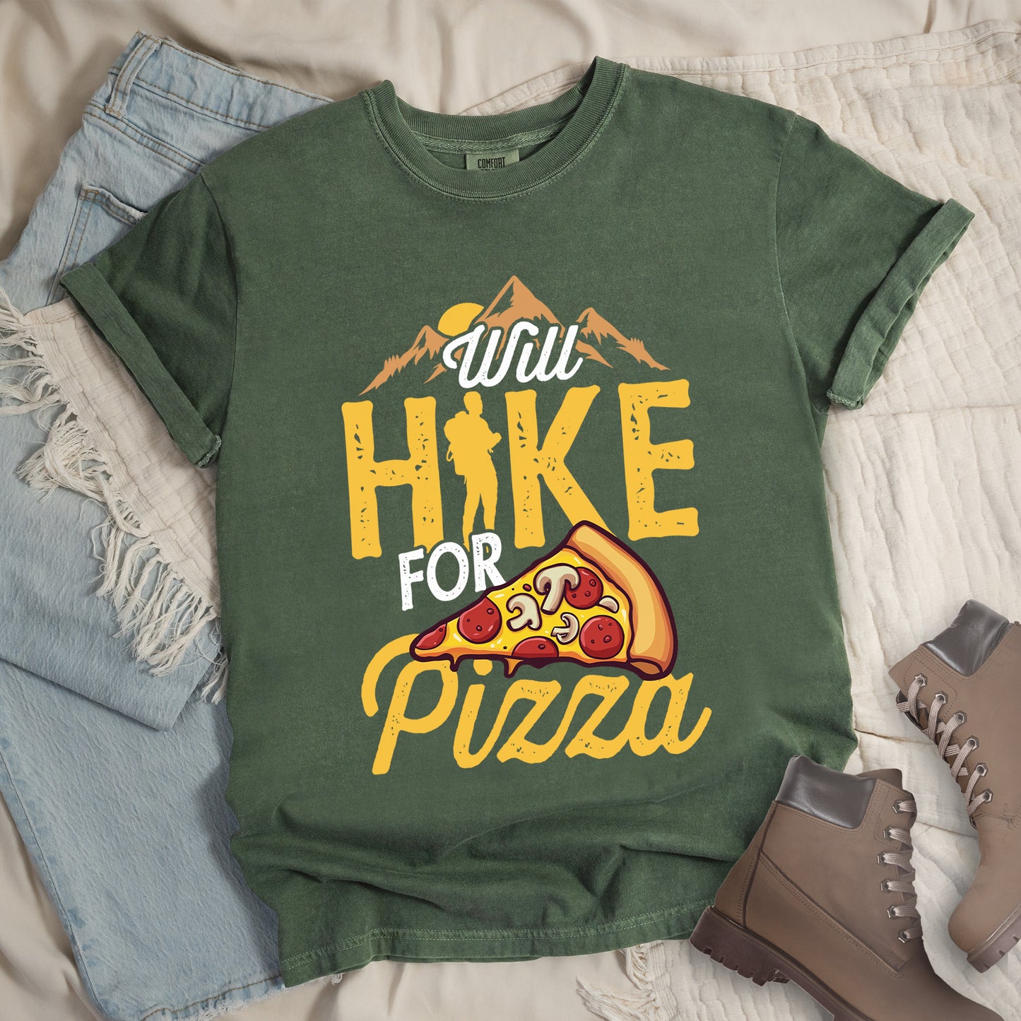 Moss(Green) t-shirt with "Will HIKE FOR pizza" design. Features "Will" in white script above mountain graphics with a yellow sun. "HIKE" appears in large yellow distressed letters with a hiker silhouette replacing the letter "I". "FOR" in white text sits above "pizza" in yellow script. An illustrated pizza slice with pepperoni and mushrooms appears between "FOR" and "pizza". The shirt features a classic crew neck and casual rolled sleeves, styled alongside light wash denim and brown hiking boots.