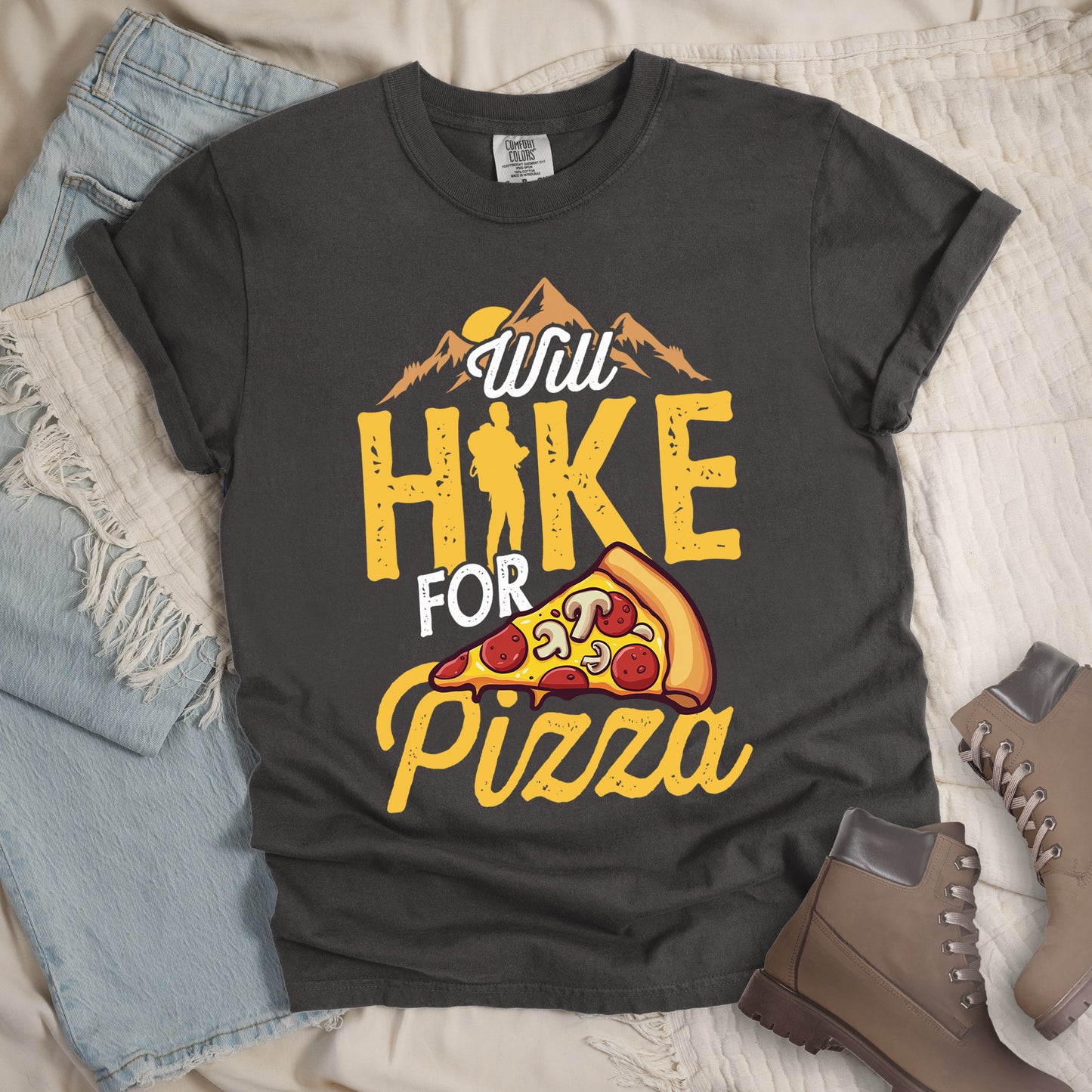 Pepper (Light Gray) t-shirt with "Will HIKE FOR pizza" design. Features "Will" in white script above mountain graphics with a yellow sun. "HIKE" appears in large yellow distressed letters with a hiker silhouette replacing the letter "I". "FOR" in white text sits above "pizza" in yellow script. An illustrated pizza slice with pepperoni and mushrooms appears between "FOR" and "pizza". The shirt features a classic crew neck and casual rolled sleeves, styled alongside light wash denim and brown hiking boots.