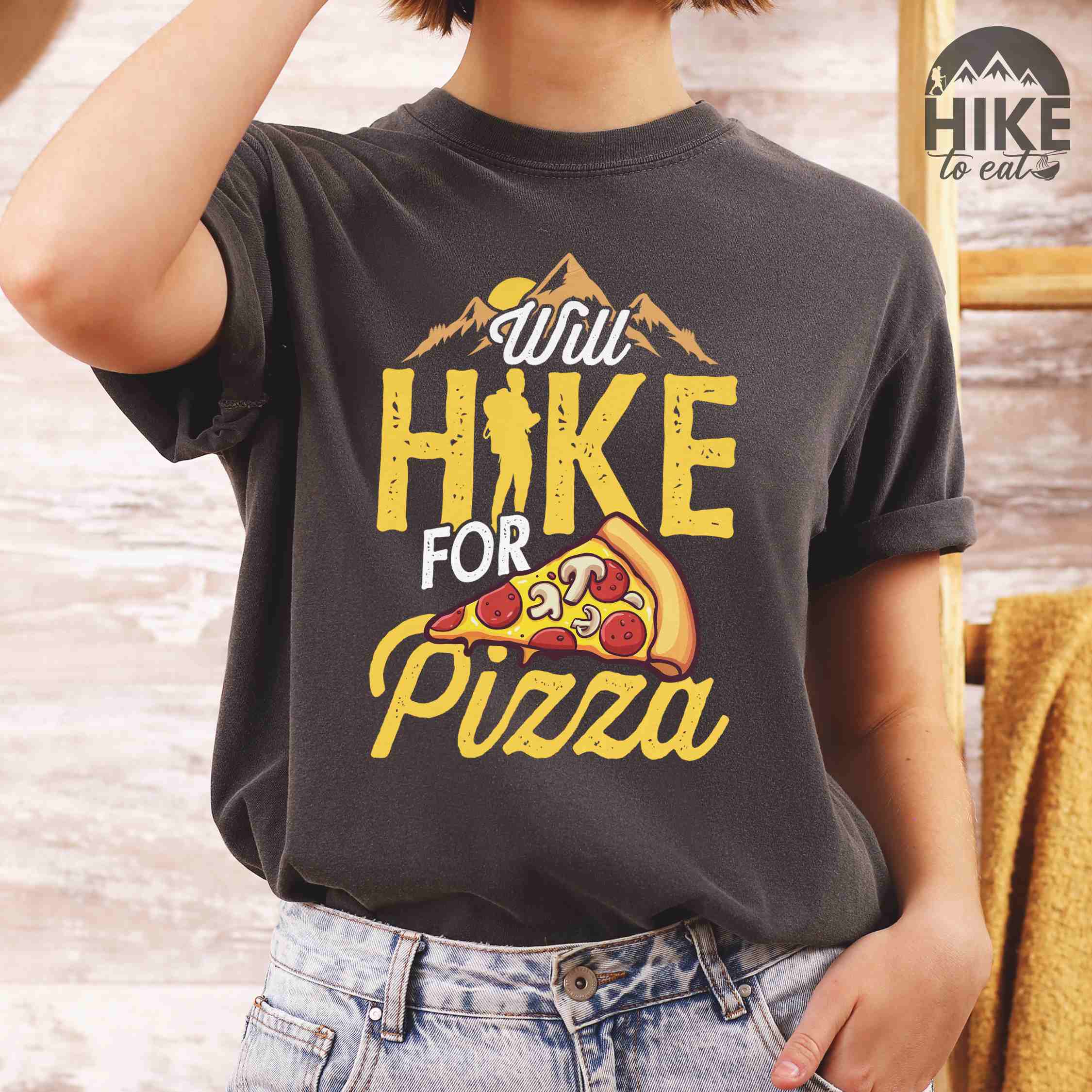 Woman wearing pepper colored vintage tee featuring Hiking design of mountains with a sunrise behind and a slice of pizza in front of the phrase 'Will Hike for Pizza'.