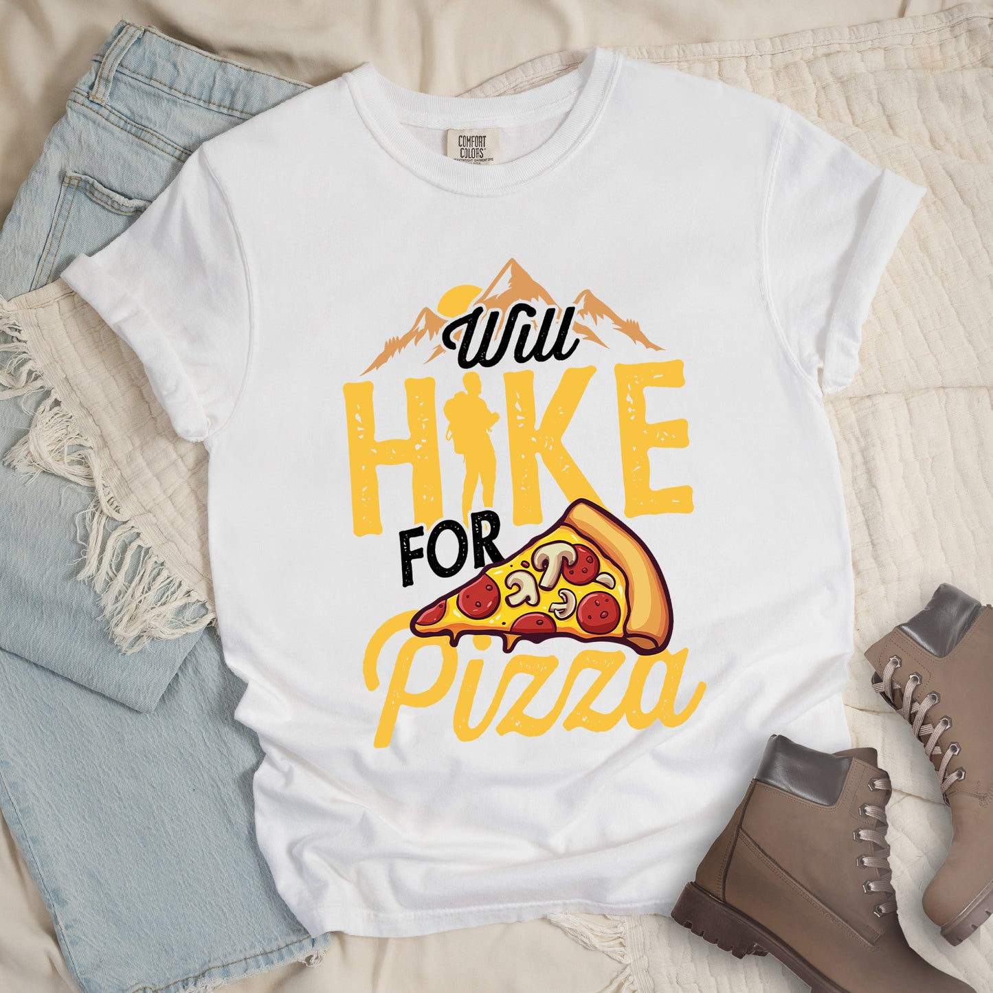 White t-shirt with "Will HIKE FOR pizza" design. Features "Will" in black script above mountain graphics with a yellow sun. "HIKE" appears in large yellow distressed letters with a hiker silhouette replacing the letter "I". "FOR" in black text sits above "pizza" in yellow script. An illustrated pizza slice with pepperoni and mushrooms appears between "FOR" and "pizza". The shirt features a classic crew neck and casual rolled sleeves, styled alongside light wash denim and brown hiking boots.