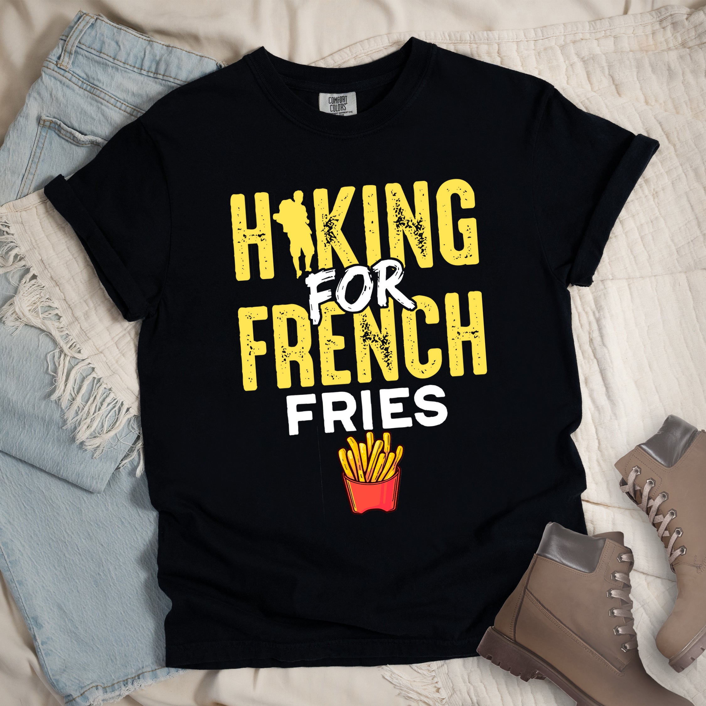 Black t-shirt with yellow and white distressed text reading "HIKING FOR FRENCH FRIES". "HIKING" appears in yellow at the top with a hiker silhouette integrated into the letter "I", "FOR" in white brushstroke text in the middle, "FRENCH" in yellow, and "FRIES" in white at the bottom. Features an illustration of french fries in a red container below the text.Features a classic crew neck and casual rolled sleeves, styled alongside light wash denim and brown hiking boots.