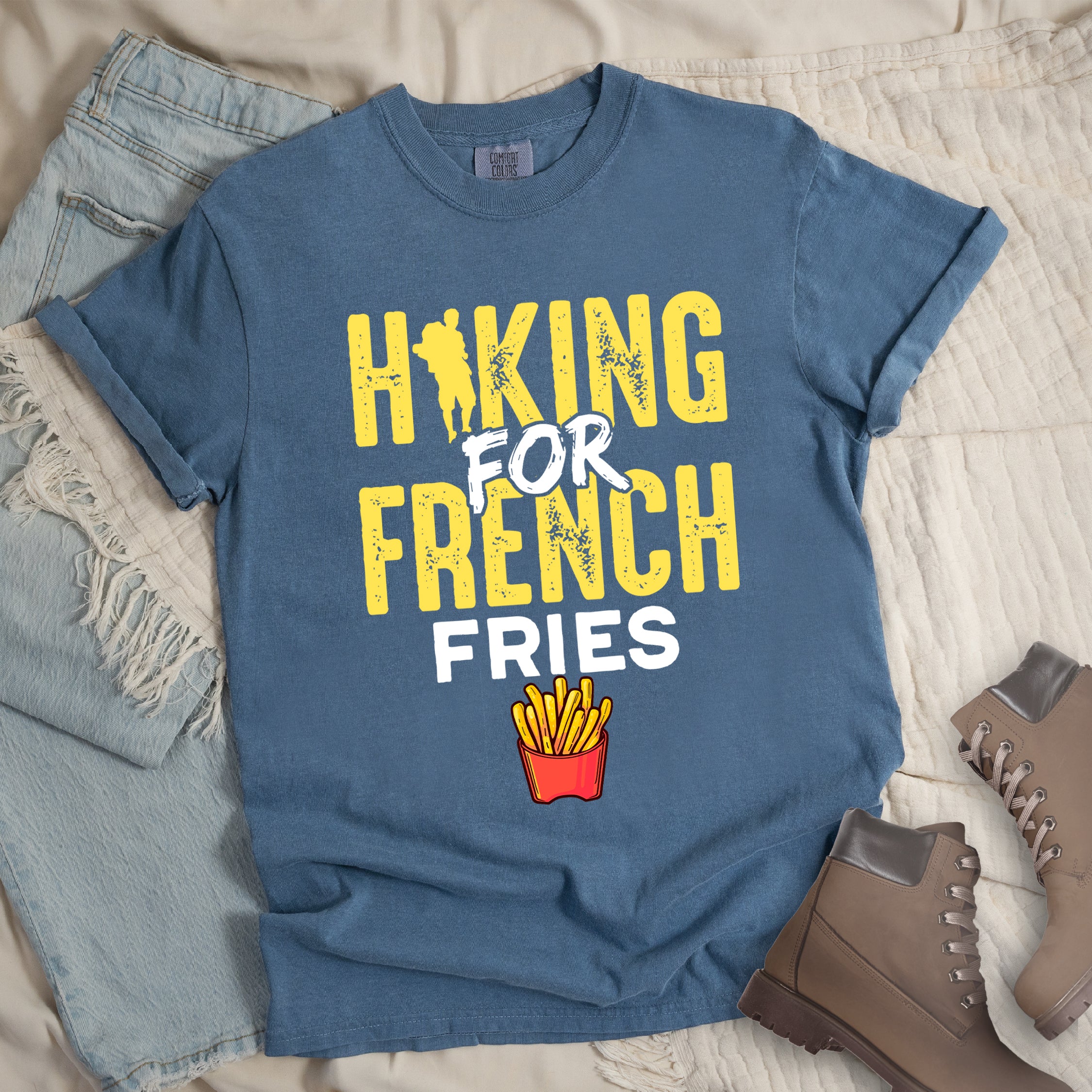 Blue Jean (Denim Blue) t-shirt with yellow and white distressed text reading "HIKING FOR FRENCH FRIES". "HIKING" appears in yellow at the top with a hiker silhouette integrated into the letter "I", "FOR" in white brushstroke text in the middle, "FRENCH" in yellow, and "FRIES" in white at the bottom. Features an illustration of french fries in a red container below the text. Features a classic crew neck and casual rolled sleeves, styled alongside light wash denim and brown hiking boots.
