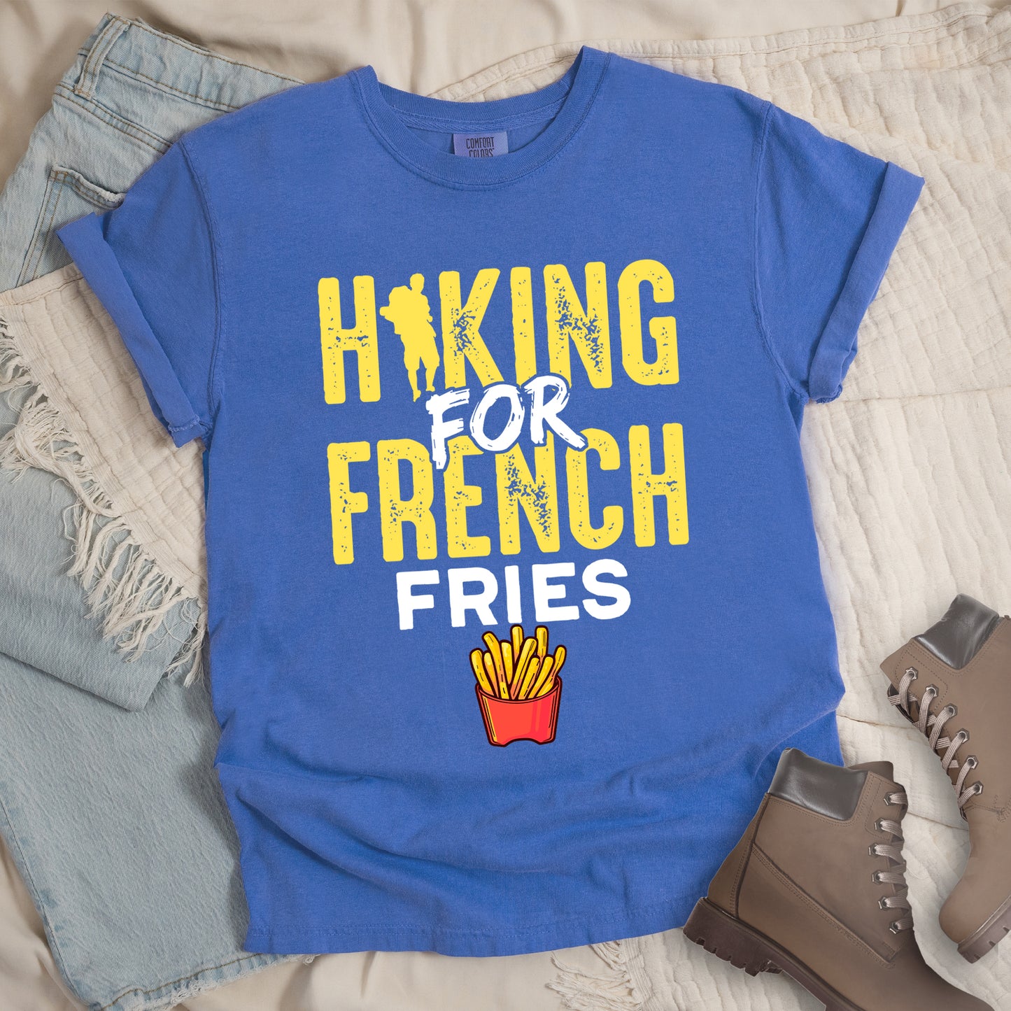 Flo Blue (Bright Blue) t-shirt with yellow and white distressed text reading "HIKING FOR FRENCH FRIES". "HIKING" appears in yellow at the top with a hiker silhouette integrated into the letter "I", "FOR" in white brushstroke text in the middle, "FRENCH" in yellow, and "FRIES" in white at the bottom. Features an illustration of french fries in a red container below the text. Features a classic crew neck and casual rolled sleeves, styled alongside light wash denim and brown hiking boots.