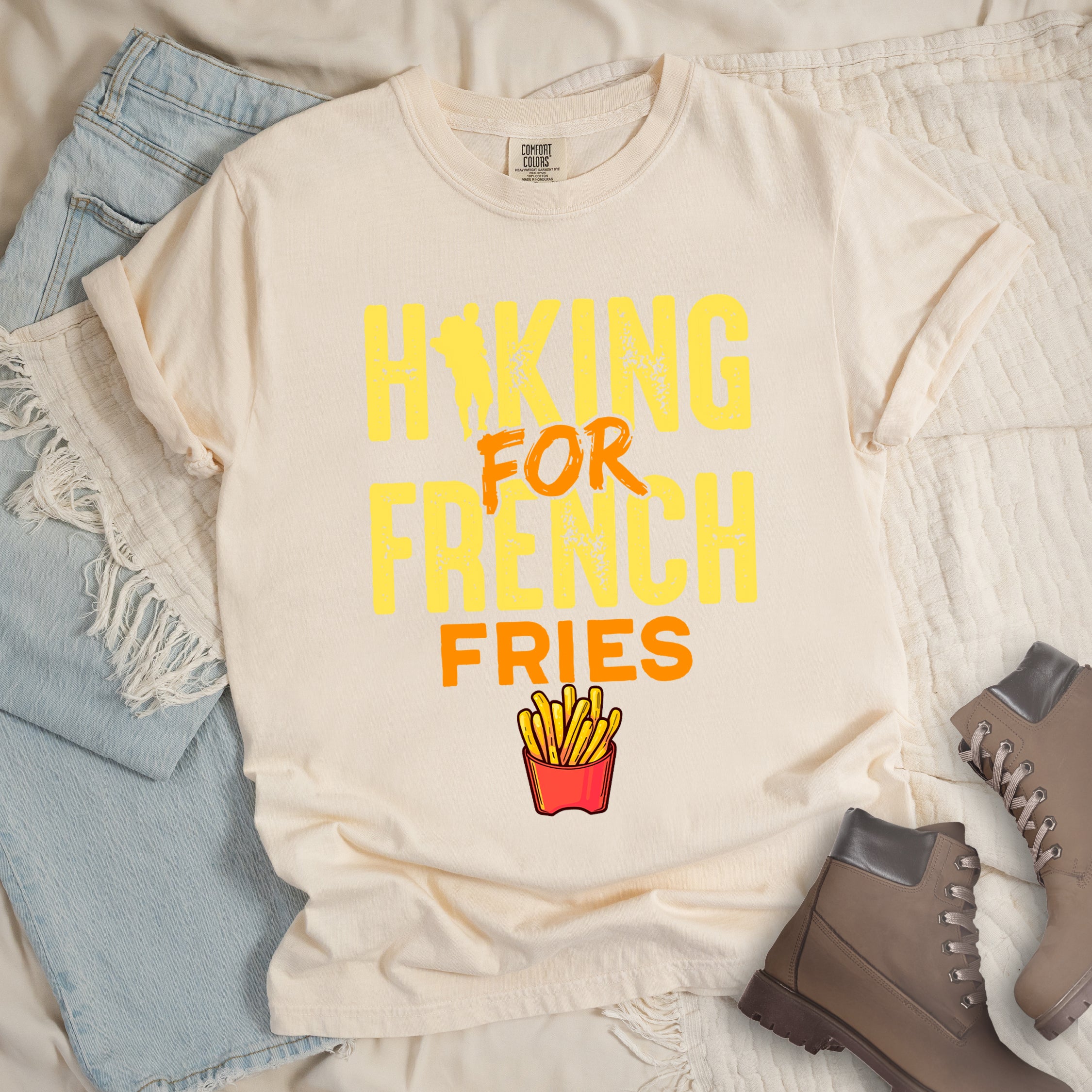 Ivory (Cream White) t-shirt with yellow and orange distressed text reading "HIKING FOR FRENCH FRIES". "HIKING" appears in yellow at the top with a hiker silhouette integrated into the letter "I", "FOR" in orange brushstroke text in the middle, "FRENCH" in yellow, and "FRIES" in orange at the bottom. Features an illustration of french fries in a red container below the text. Features a classic crew neck and casual rolled sleeves, styled alongside light wash denim and brown hiking boots.