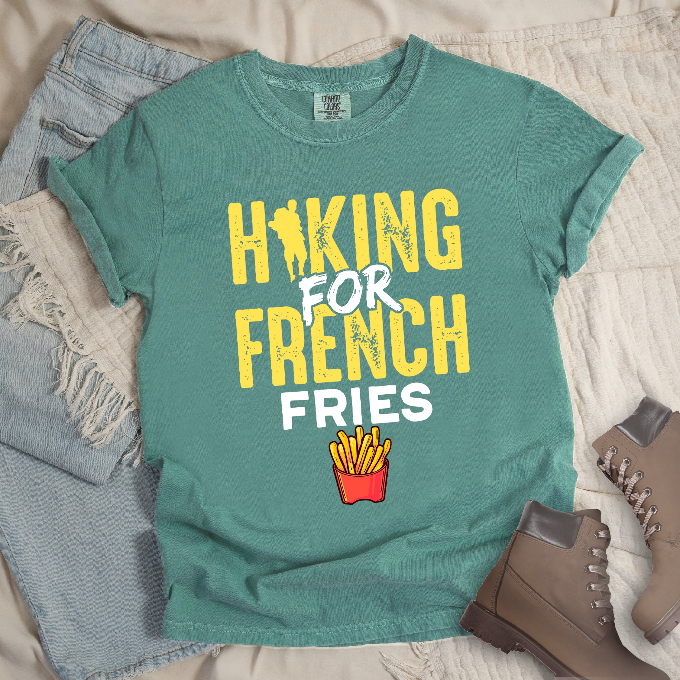 Light Green t-shirt with yellow and white distressed text reading "HIKING FOR FRENCH FRIES". "HIKING" appears in yellow at the top with a hiker silhouette integrated into the letter "I", "FOR" in white brushstroke text in the middle, "FRENCH" in yellow, and "FRIES" in white at the bottom. Features an illustration of french fries in a red container below the text. Features a classic crew neck and casual rolled sleeves, styled alongside light wash denim and brown hiking boots.