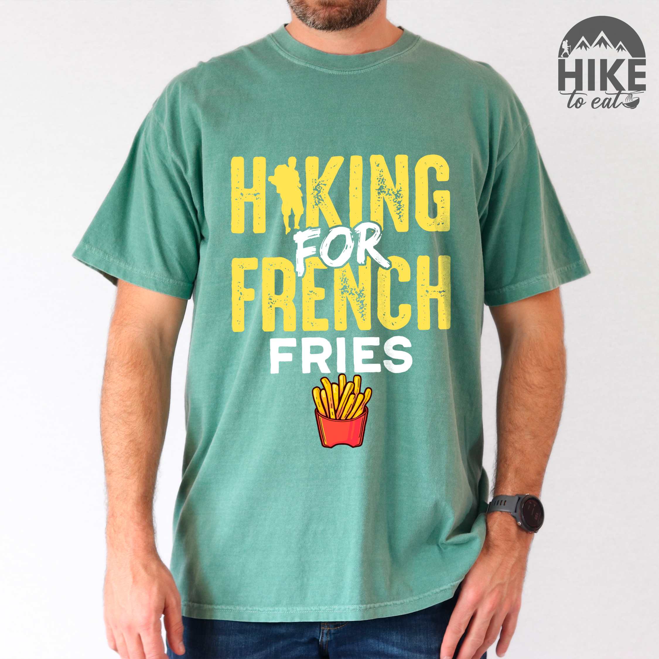 Man wearing light green "Hiking for french fries" colored vintage tee with bold typography, mountains, illustrated fries, and backpacker for comfort food enthusiasts.