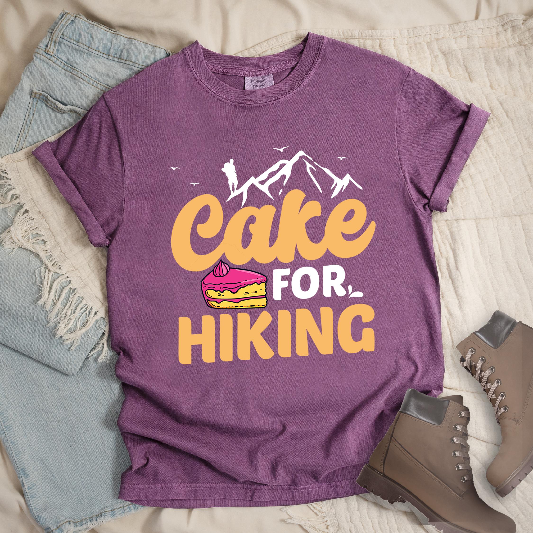Dark pink t-shirt with text reading "Cake FOR HIKING". Features white mountain silhouettes with a hiker figure and birds at the top, and an illustration of a yellow cake slice with pink frosting between the words.