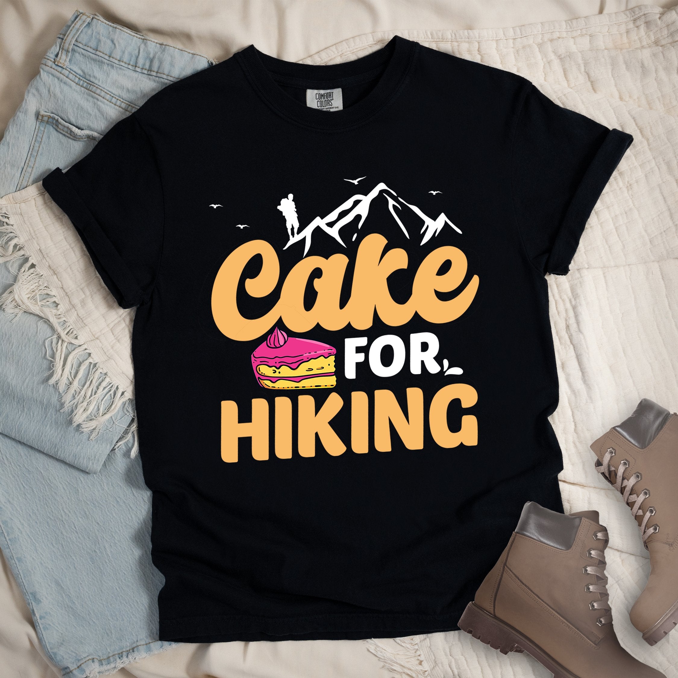 Black t-shirt with text reading "Cake FOR HIKING". Features white mountain silhouettes with a hiker figure and birds at the top, and an illustration of a yellow cake slice with pink frosting between the words.