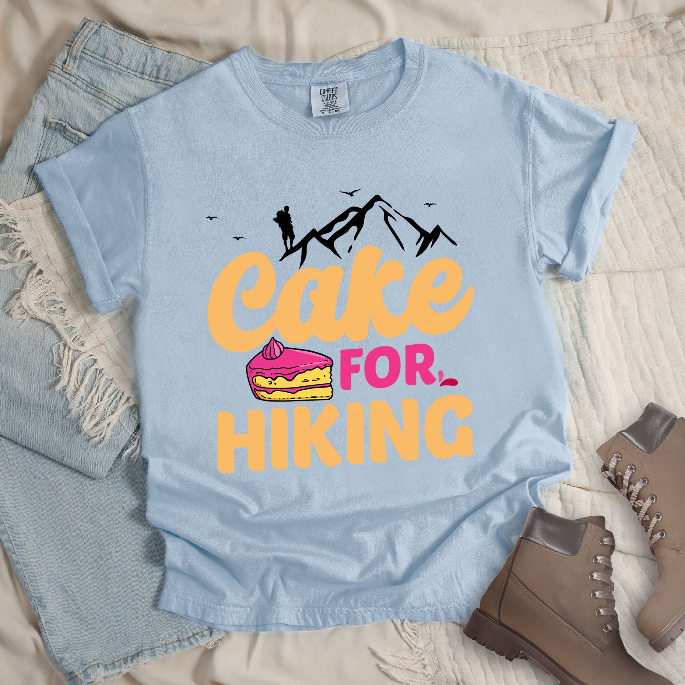 Light blue t-shirt with text reading "Cake FOR HIKING". Features white mountain silhouettes with a hiker figure and birds at the top, and an illustration of a yellow cake slice with pink frosting between the words.