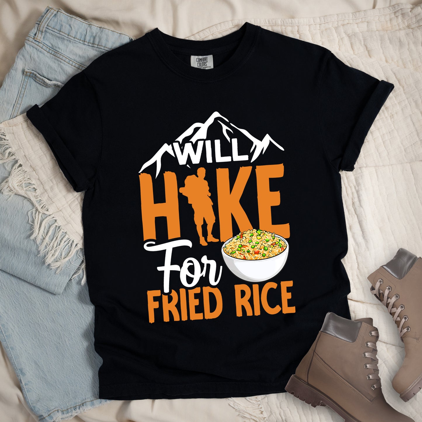 Black t-shirt with white, orange and white text reading "WILL HIKE For FRIED RICE". "WILL" appears in white over mountain silhouettes at the top, "HIKE" in large orange letters with a hiker silhouette integrated into the letter "I", "For" in white script in the middle, and "FRIED RICE" in orange at the bottom. Features an illustration of a white bowl filled with colorful fried rice. Features a classic crew neck and casual rolled sleeves, styled alongside light wash denim and brown hiking boots.