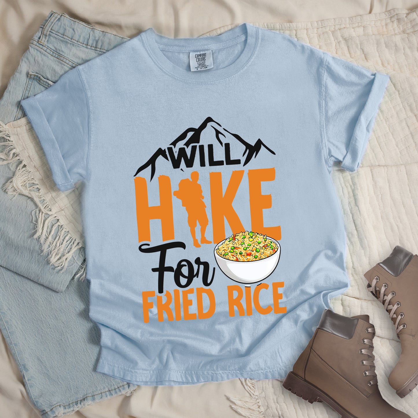 Chambray (Light Blue) t-shirt with white, orange and white text reading "WILL HIKE For FRIED RICE". "WILL" appears in black over mountain silhouettes at the top, "HIKE" in large orange letters with a hiker silhouette integrated into the letter "I", "For" in black script in the middle, and "FRIED RICE" in orange at the bottom. Features an illustration of a white bowl filled with colorful fried rice. Features a classic crew neck and casual rolled sleeves, alongside light wash denim and brown hiking boots.