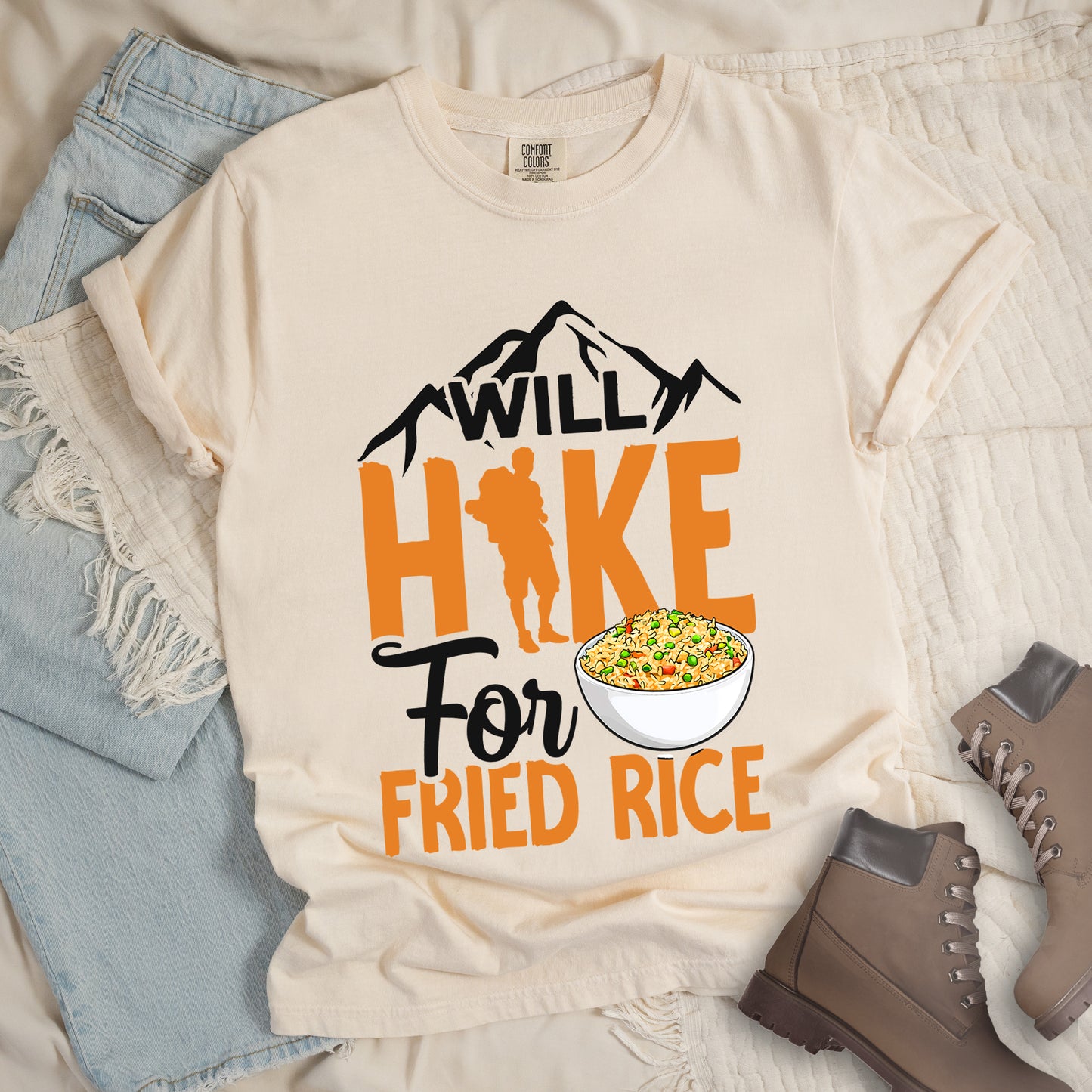 Ivory (Cream White) t-shirt with black, and orange text reading "WILL HIKE For FRIED RICE". "WILL" appears in black over mountain silhouettes at the top, "HIKE" in large orange letters with a hiker silhouette integrated into the letter "I", "For" in black script in the middle, and "FRIED RICE" in orange at the bottom. Features an illustration of a white bowl filled with colorful fried rice. Features a classic crew neck and casual rolled sleeves, styled alongside light wash denim and brown hiking boots.