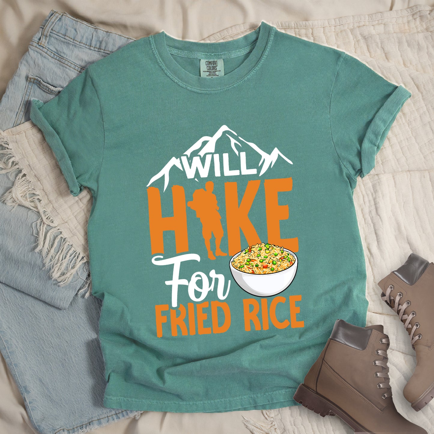 Light Green t-shirt with white, orange and white text reading "WILL HIKE For FRIED RICE". "WILL" appears in white over mountain silhouettes at the top, "HIKE" in large orange letters with a hiker silhouette integrated into the letter "I", "For" in white script in the middle, and "FRIED RICE" in orange at the bottom. Features an illustration of a white bowl filled with colorful fried rice. Features a classic crew neck and casual rolled sleeves, styled alongside light wash denim and brown hiking boots.