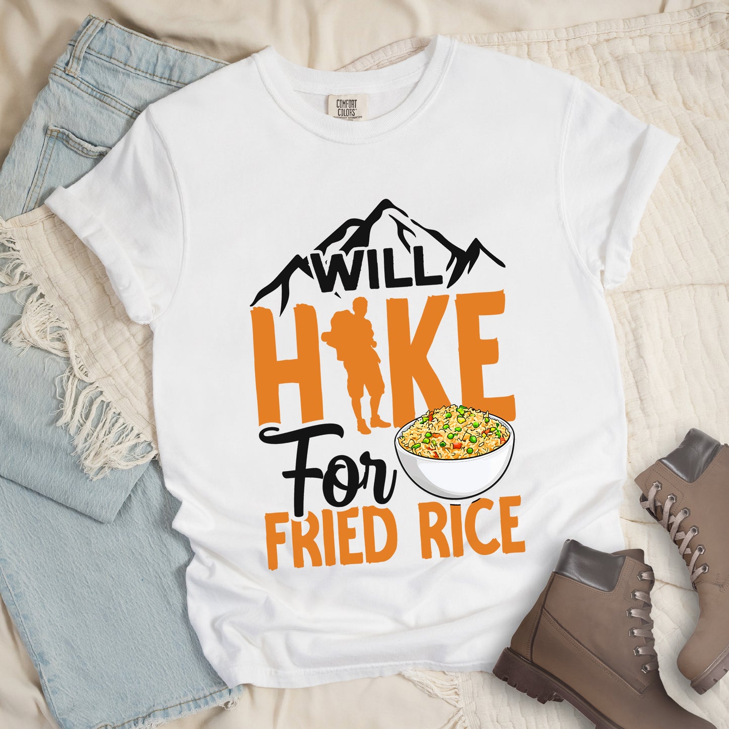White t-shirt with black, and orange text reading "WILL HIKE For FRIED RICE". "WILL" appears in black over mountain silhouettes at the top, "HIKE" in large orange letters with a hiker silhouette integrated into the letter "I", "For" in black script in the middle, and "FRIED RICE" in orange at the bottom. Features an illustration of a white bowl filled with colorful fried rice.Features a classic crew neck and casual rolled sleeves, styled alongside light wash denim and brown hiking boots.