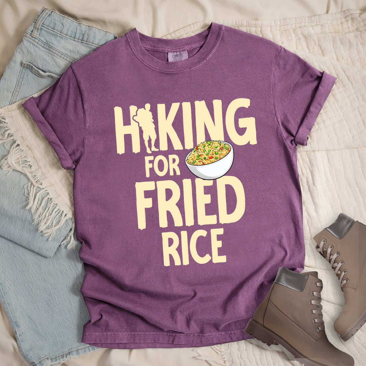 Berry (Dark Violet) t-shirt with pale yellow text reading "HIKING FOR FRIED RICE". "HIKING" appears at the top with a hiker silhouette integrated into the letter "I", "FOR" in smaller text in the middle, and "FRIED RICE" in large letters at the bottom. Features an illustration of a white bowl filled with colorful fried rice with vegetables next to "FOR" and above "FRIED". Features a classic crew neck and casual rolled sleeves, styled alongside light wash denim and brown hiking boots.