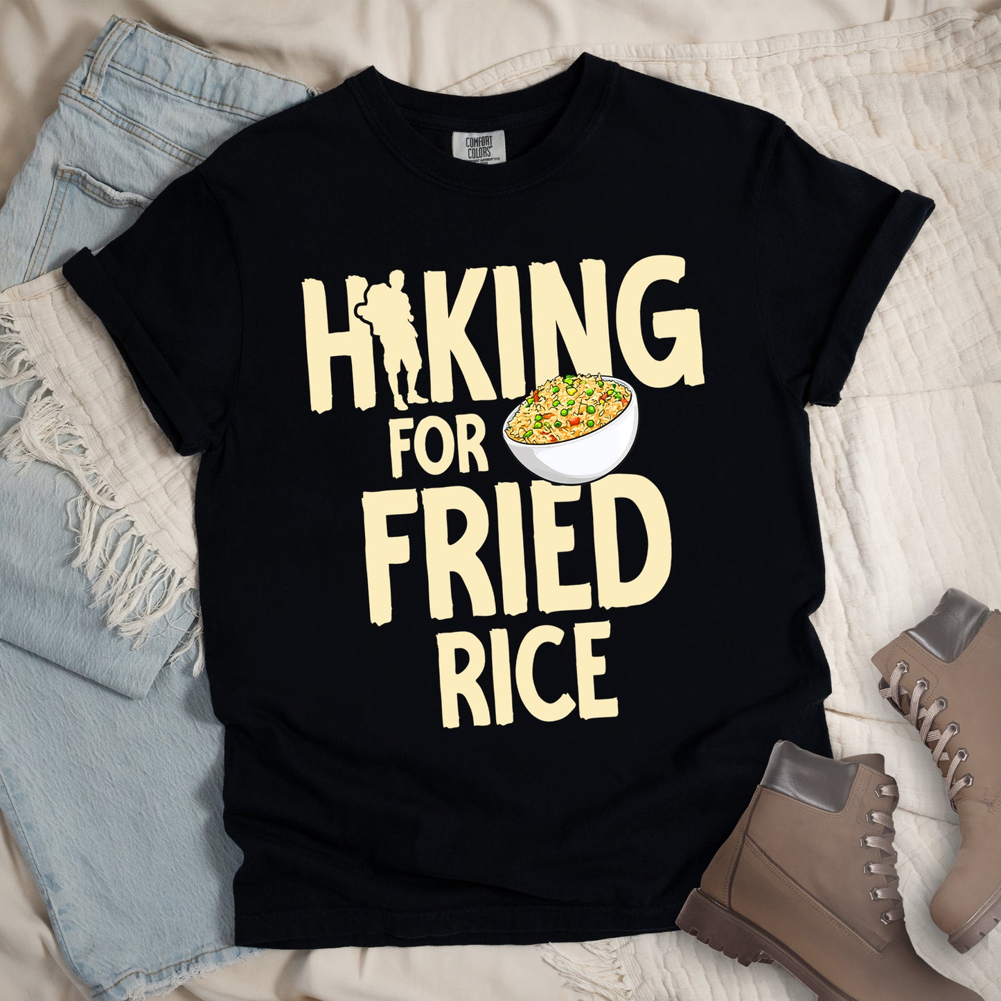 Black t-shirt with pale yellow text reading "HIKING FOR FRIED RICE". "HIKING" appears at the top with a hiker silhouette integrated into the letter "I", "FOR" in smaller text in the middle, and "FRIED RICE" in large letters at the bottom. Features an illustration of a white bowl filled with colorful fried rice with vegetables next to "FOR" and above "FRIED". Features a classic crew neck and casual rolled sleeves, styled alongside light wash denim and brown hiking boots.