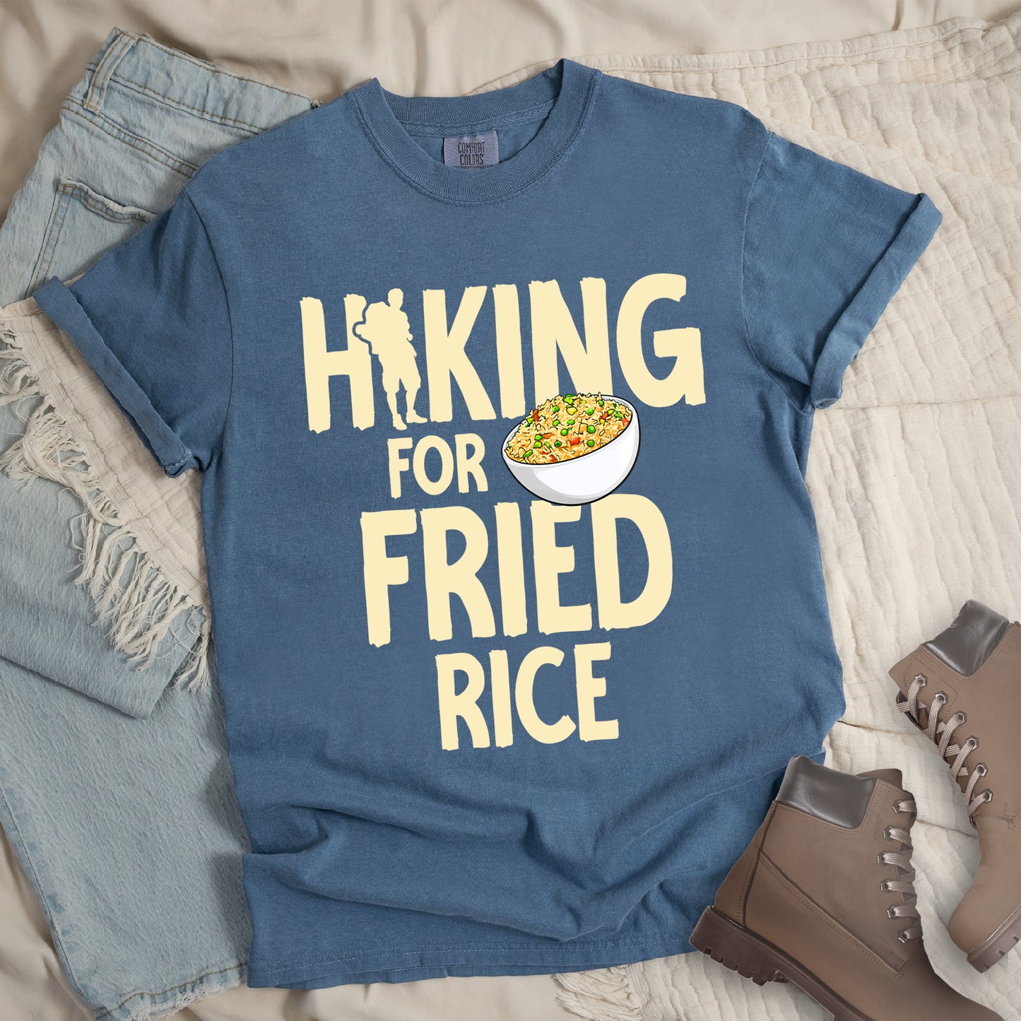 Blue Jean (Denim Blue) t-shirt with pale yellow text reading "HIKING FOR FRIED RICE". "HIKING" appears at the top with a hiker silhouette integrated into the letter "I", "FOR" in smaller text in the middle, and "FRIED RICE" in large letters at the bottom. Features an illustration of a white bowl filled with colorful fried rice with vegetables next to "FOR" and above "FRIED". Features a classic crew neck and casual rolled sleeves, styled alongside light wash denim and brown hiking boots.
