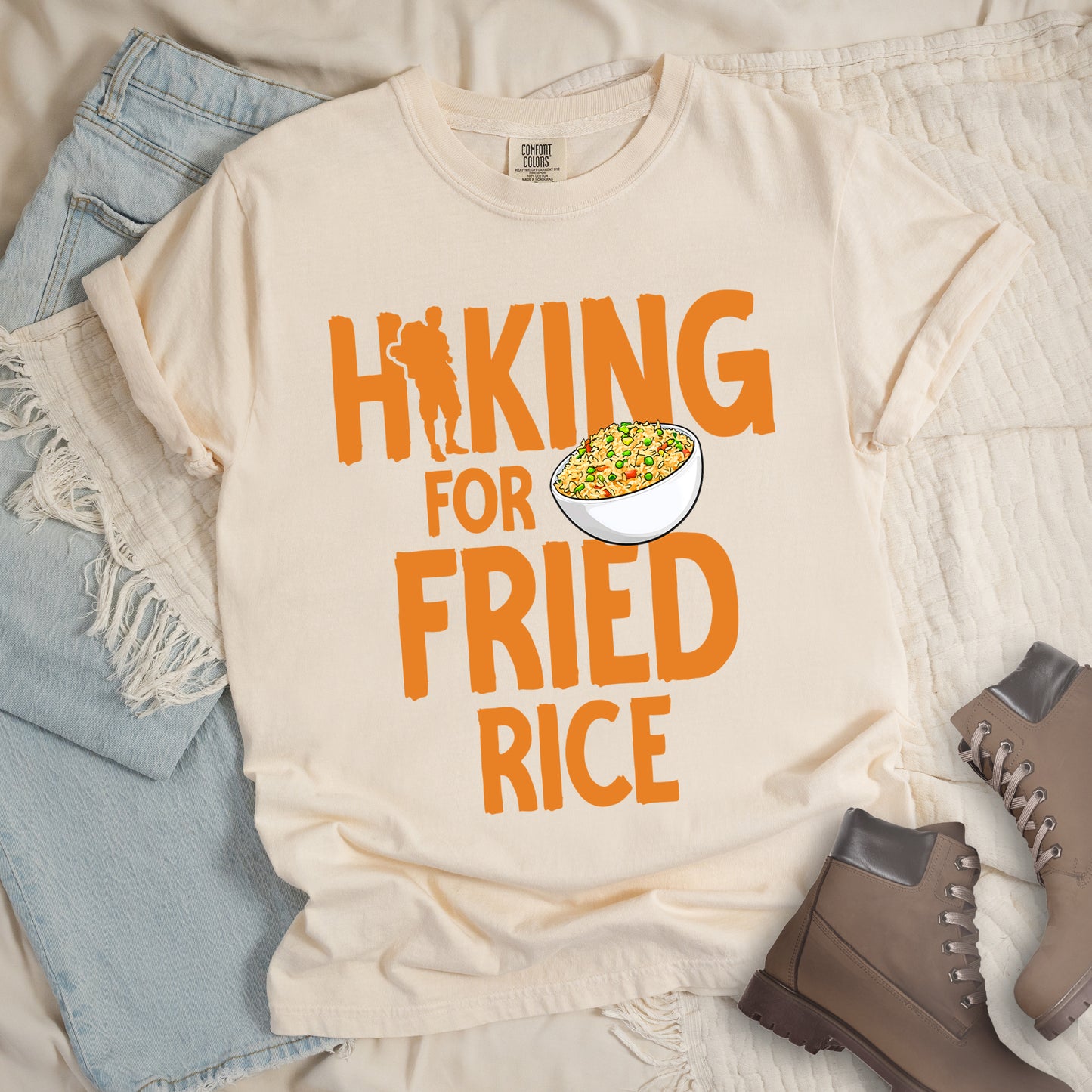 Ivory (Cream White) t-shirt with orange text reading "HIKING FOR FRIED RICE". "HIKING" appears at the top with a hiker silhouette integrated into the letter "I", "FOR" in smaller text in the middle, and "FRIED RICE" in large letters at the bottom. Features an illustration of a white bowl filled with colorful fried rice with vegetables next to "FOR" and above "FRIED". Features a classic crew neck and casual rolled sleeves, styled alongside light wash denim and brown hiking boots.