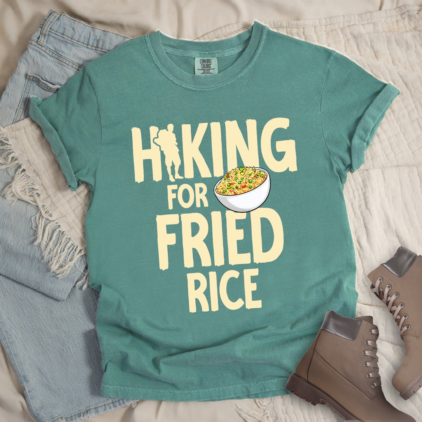Light Green t-shirt with pale yellow text reading "HIKING FOR FRIED RICE". "HIKING" appears at the top with a hiker silhouette integrated into the letter "I", "FOR" in smaller text in the middle, and "FRIED RICE" in large letters at the bottom. Features an illustration of a white bowl filled with colorful fried rice with vegetables next to "FOR" and above "FRIED". Features a classic crew neck and casual rolled sleeves, styled alongside light wash denim and brown hiking boots.