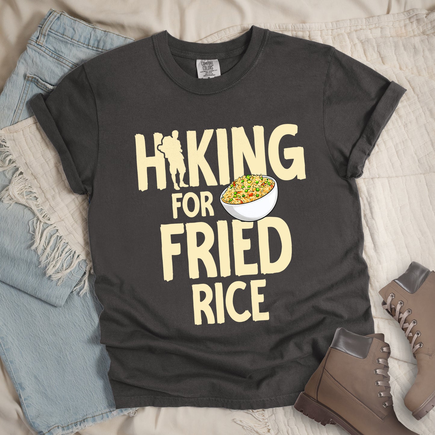 Pepper (Dark Gray) t-shirt with pale yellow text reading "HIKING FOR FRIED RICE". "HIKING" appears at the top with a hiker silhouette integrated into the letter "I", "FOR" in smaller text in the middle, and "FRIED RICE" in large letters at the bottom. Features an illustration of a white bowl filled with colorful fried rice with vegetables next to "FOR" and above "FRIED". Features a classic crew neck and casual rolled sleeves, styled alongside light wash denim and brown hiking boots.