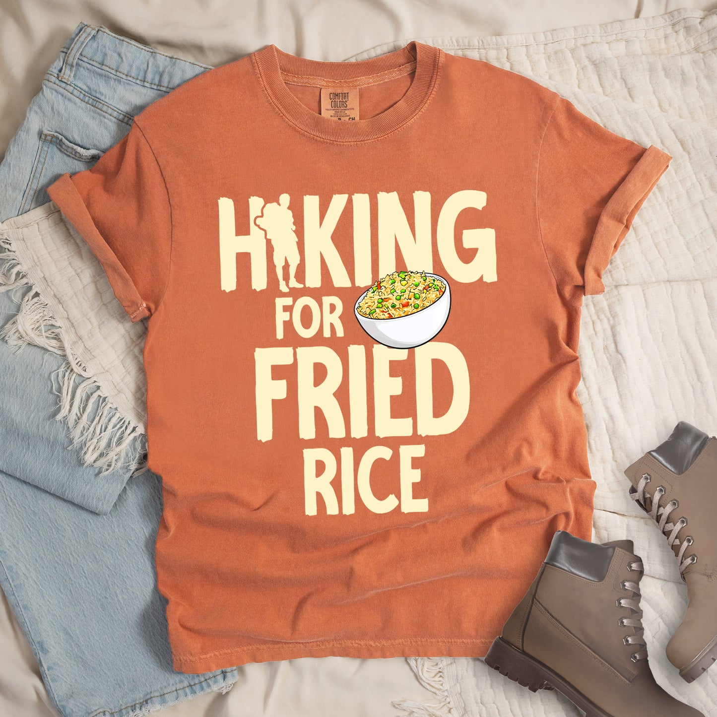 Yam (brown) t-shirt with pale yellow text reading "HIKING FOR FRIED RICE". "HIKING" appears at the top with a hiker silhouette integrated into the letter "I", "FOR" in smaller text in the middle, and "FRIED RICE" in large letters at the bottom. Features an illustration of a white bowl filled with colorful fried rice with vegetables next to "FOR" and above "FRIED". Features a classic crew neck and casual rolled sleeves, styled alongside light wash denim and brown hiking boots.