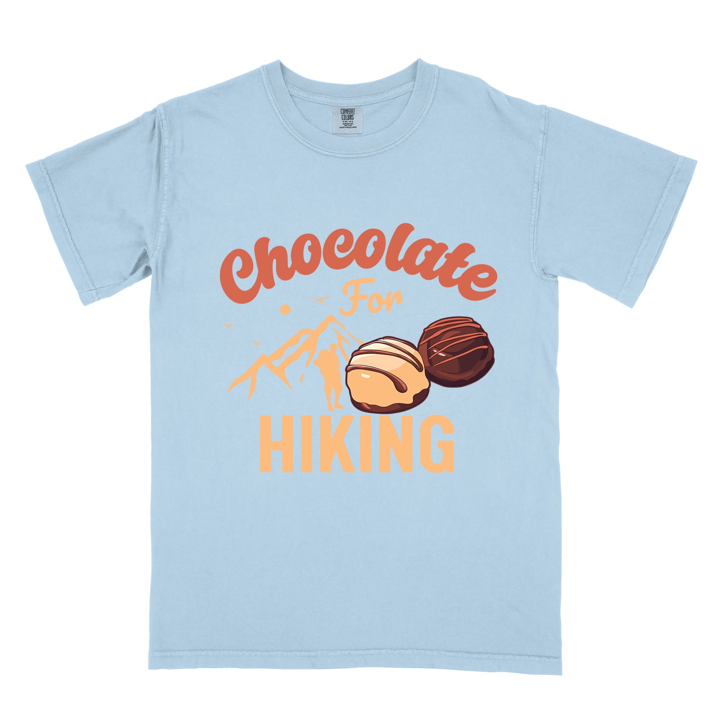 Chambray (Light Blue) t-shirt with coral and peach text reading "Chocolate For HIKING". "Chocolate" appears in curved coral script at the top, "For" in small peach script in the middle, and "HIKING" in large peach letters at the bottom. Features illustrations of two chocolate truffles (one light, one dark) in the center, peach mountain silhouettes with small birds, and a hiker figure silhouette.