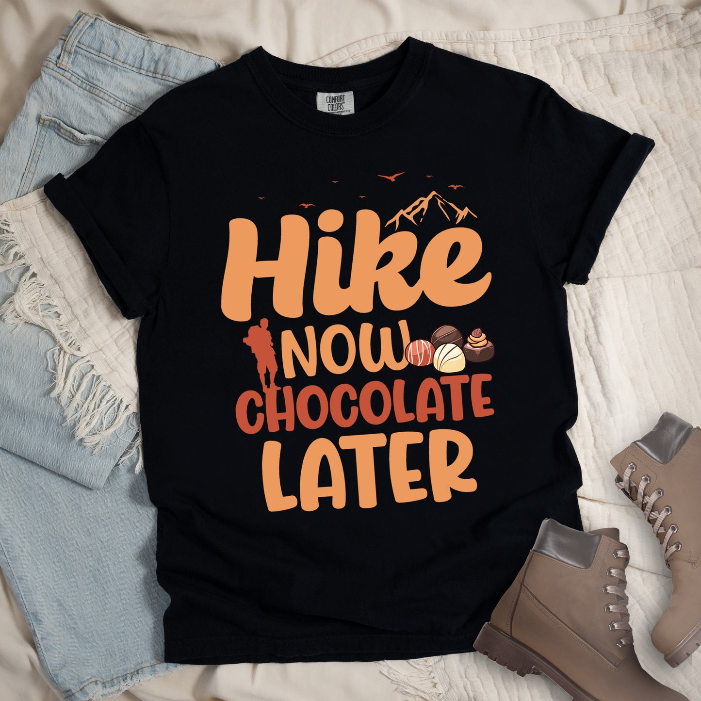 Black t-shirt with orange and brown text reading "Hike NOW CHOCOLATE LATER". "Hike" appears in large orange at the top with small orange mountain silhouettes and bird silhouettes, "NOW" in lighter orange with a red hiker silhouette, "CHOCOLATE" in brown, and "LATER" in orange at the bottom. Features illustrations of various chocolate truffles between "NOW" and "CHOCOLATE". Features a classic crew neck and casual rolled sleeves, styled alongside light wash denim and brown hiking boots.