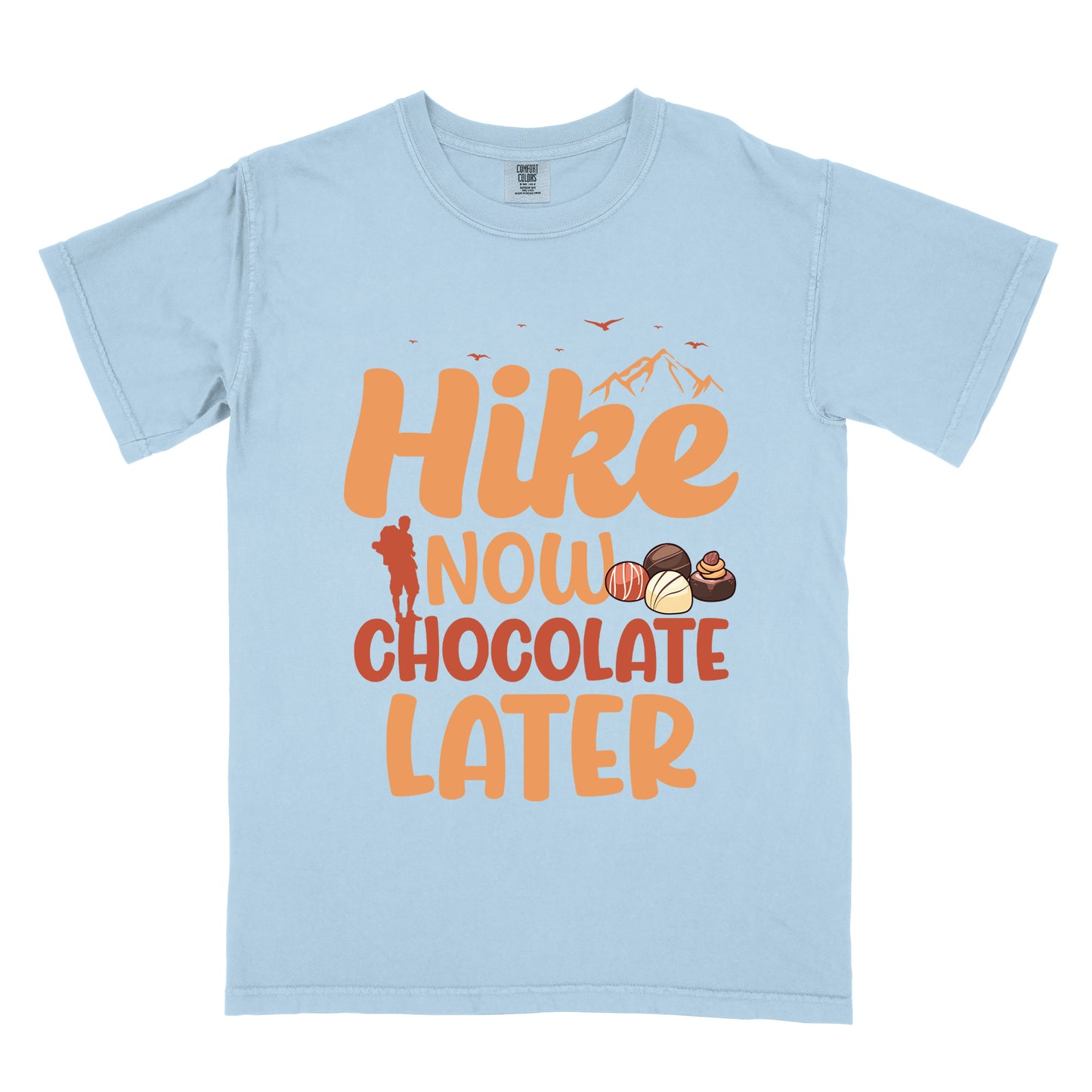 Chambray (Light Blue) t-shirt with orange and brown text reading "Hike NOW CHOCOLATE LATER". "Hike" appears in large orange at the top with small orange mountain silhouettes and bird silhouettes, "NOW" in lighter orange with a red hiker silhouette, "CHOCOLATE" in brown, and "LATER" in orange at the bottom. Features illustrations of various chocolate truffles between "NOW" and "CHOCOLATE".
