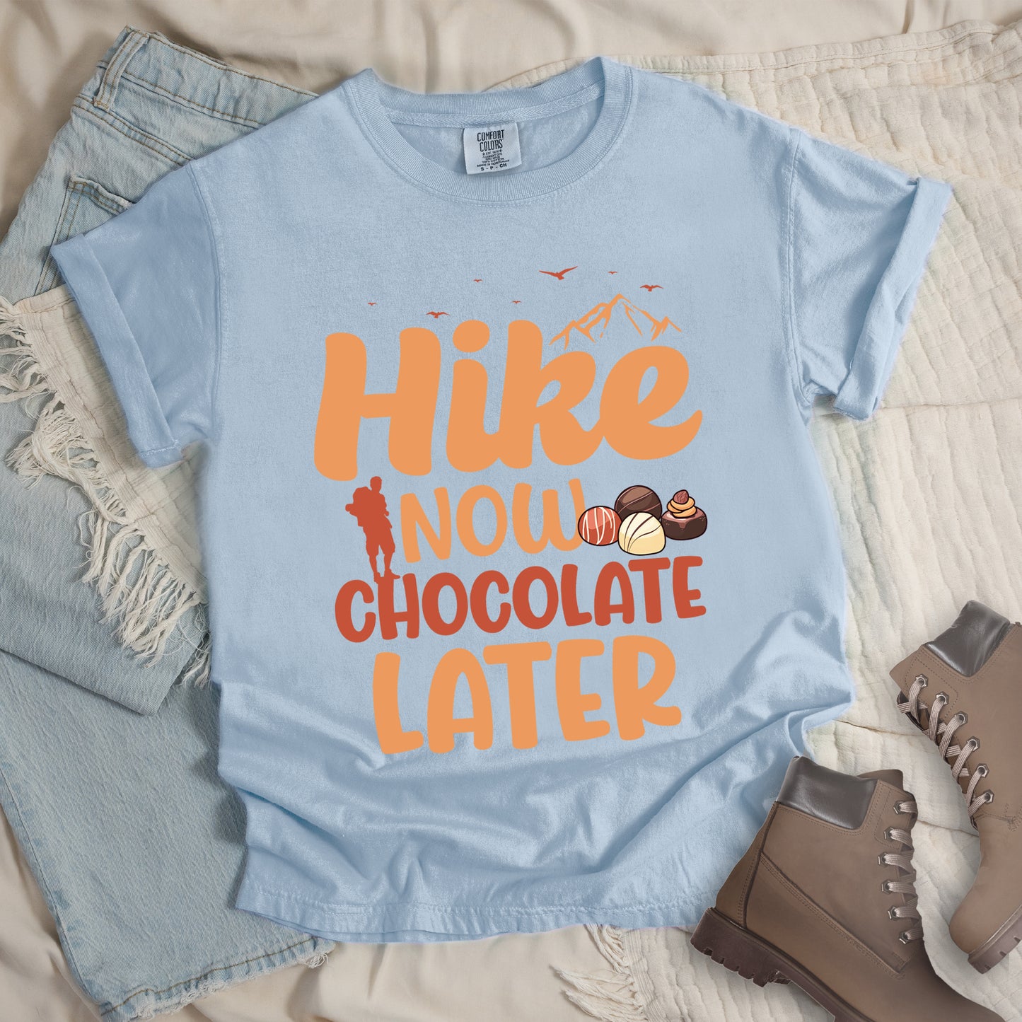 Chambray (Light Blue) t-shirt with orange and brown text reading "Hike NOW CHOCOLATE LATER". "Hike" appears in large orange at the top with small orange mountain silhouettes and bird silhouettes, "NOW" in lighter orange with a red hiker silhouette, "CHOCOLATE" in brown, and "LATER" in orange at the bottom. Features illustrations of various chocolate truffles between "NOW" and "CHOCOLATE". Features a classic crew neck and casual rolled sleeves, styled alongside light wash denim and brown hiking boots.
