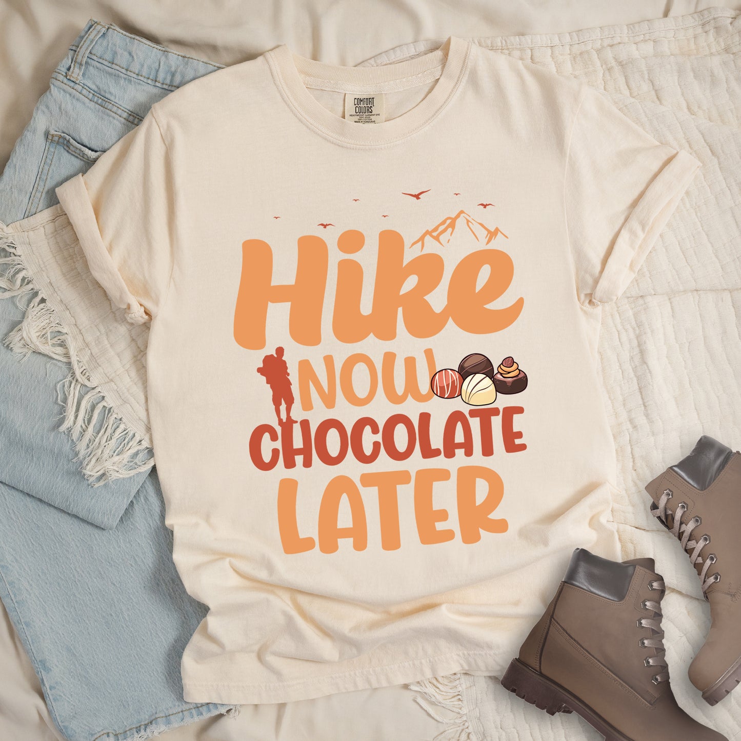 Ivory (Cream White) t-shirt with orange and brown text reading "Hike NOW CHOCOLATE LATER". "Hike" appears in large orange at the top with small orange mountain silhouettes and bird silhouettes, "NOW" in lighter orange with a red hiker silhouette, "CHOCOLATE" in brown, and "LATER" in orange at the bottom. Features illustrations of various chocolate truffles between "NOW" and "CHOCOLATE". Features a classic crew neck and casual rolled sleeves, styled alongside light wash denim and brown hiking boots.