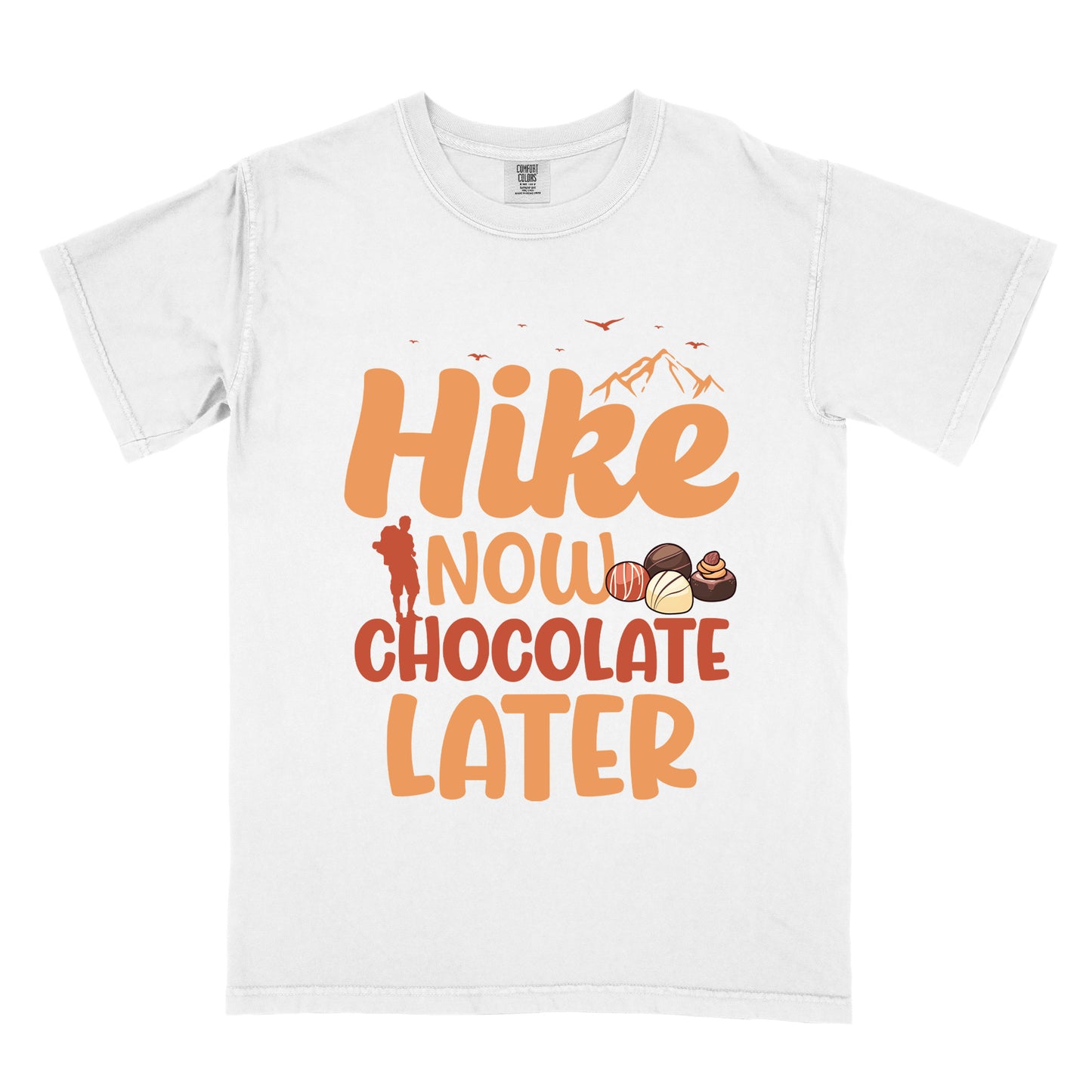 White t-shirt with orange and brown text reading "Hike NOW CHOCOLATE LATER". "Hike" appears in large orange at the top with small orange mountain silhouettes and bird silhouettes, "NOW" in lighter orange with a red hiker silhouette, "CHOCOLATE" in brown, and "LATER" in orange at the bottom. Features illustrations of various chocolate truffles between "NOW" and "CHOCOLATE".