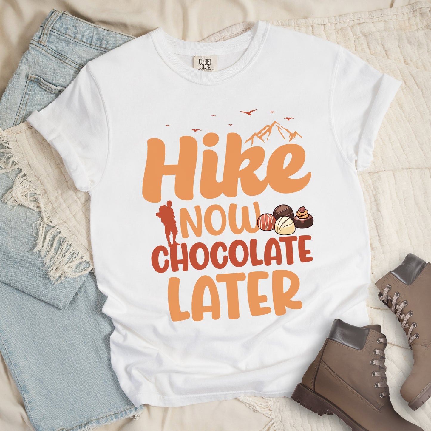 White t-shirt with orange and brown text reading "Hike NOW CHOCOLATE LATER". "Hike" appears in large orange at the top with small orange mountain silhouettes and bird silhouettes, "NOW" in lighter orange with a red hiker silhouette, "CHOCOLATE" in brown, and "LATER" in orange at the bottom. Features illustrations of various chocolate truffles between "NOW" and "CHOCOLATE". Features a classic crew neck and casual rolled sleeves, styled alongside light wash denim and brown hiking boots.