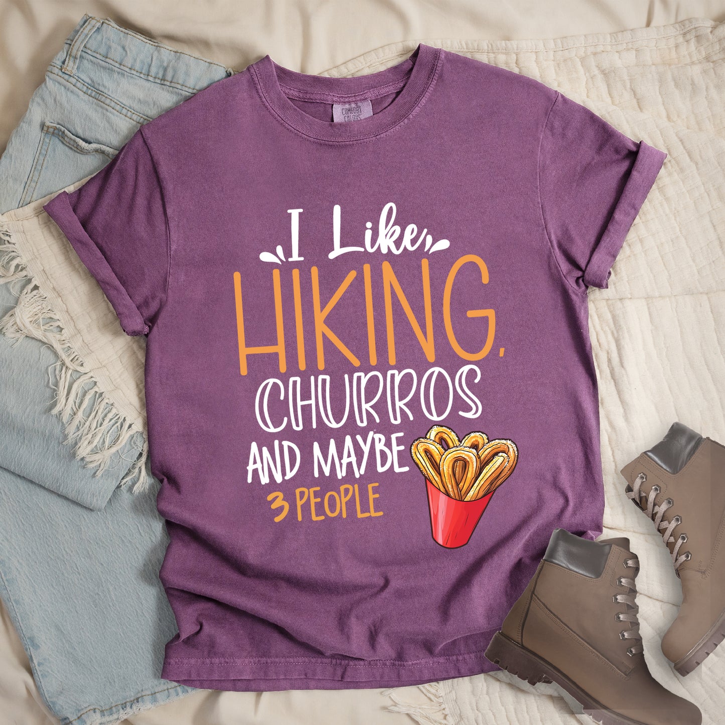 Berry (Dark Violet) t-shirt with white and orange text reading "I Like HIKING, CHURROS AND MAYBE 3 PEOPLE". "I Like" appears in white script at the top, "HIKING," in large orange letters, "CHURROS" in white outlined letters, "AND MAYBE" in white, and "3 PEOPLE" in small orange text at the bottom. Features an illustration of churros in a red container on the right side. Features a classic crew neck and casual rolled sleeves, styled alongside light wash denim and brown hiking boots.