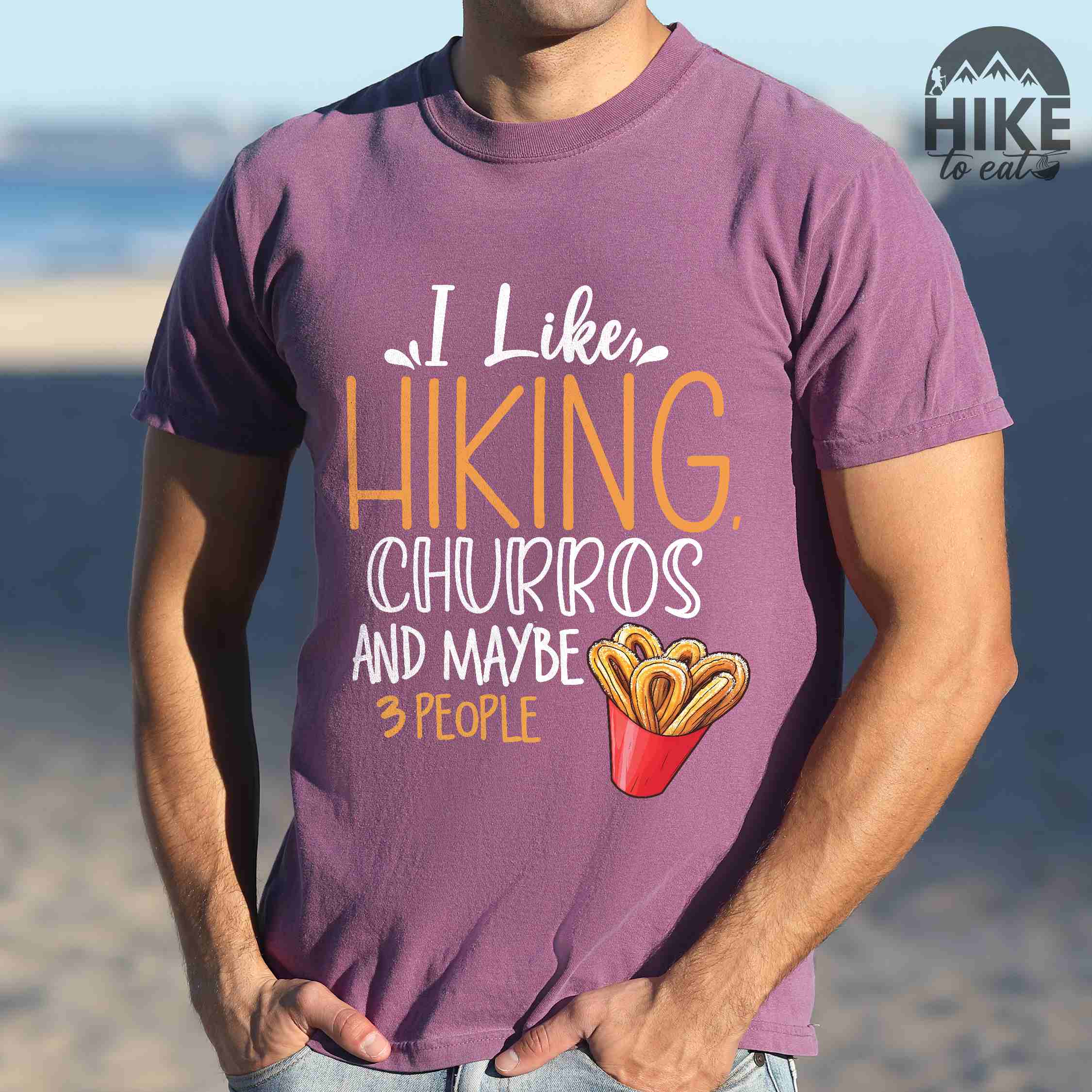 Man wearing berry "I like hiking, churros and maybe three people " colored vintage tee with bold typography, mountains, illustrated a serving of Churros, and backpackers with a sweet tooth.