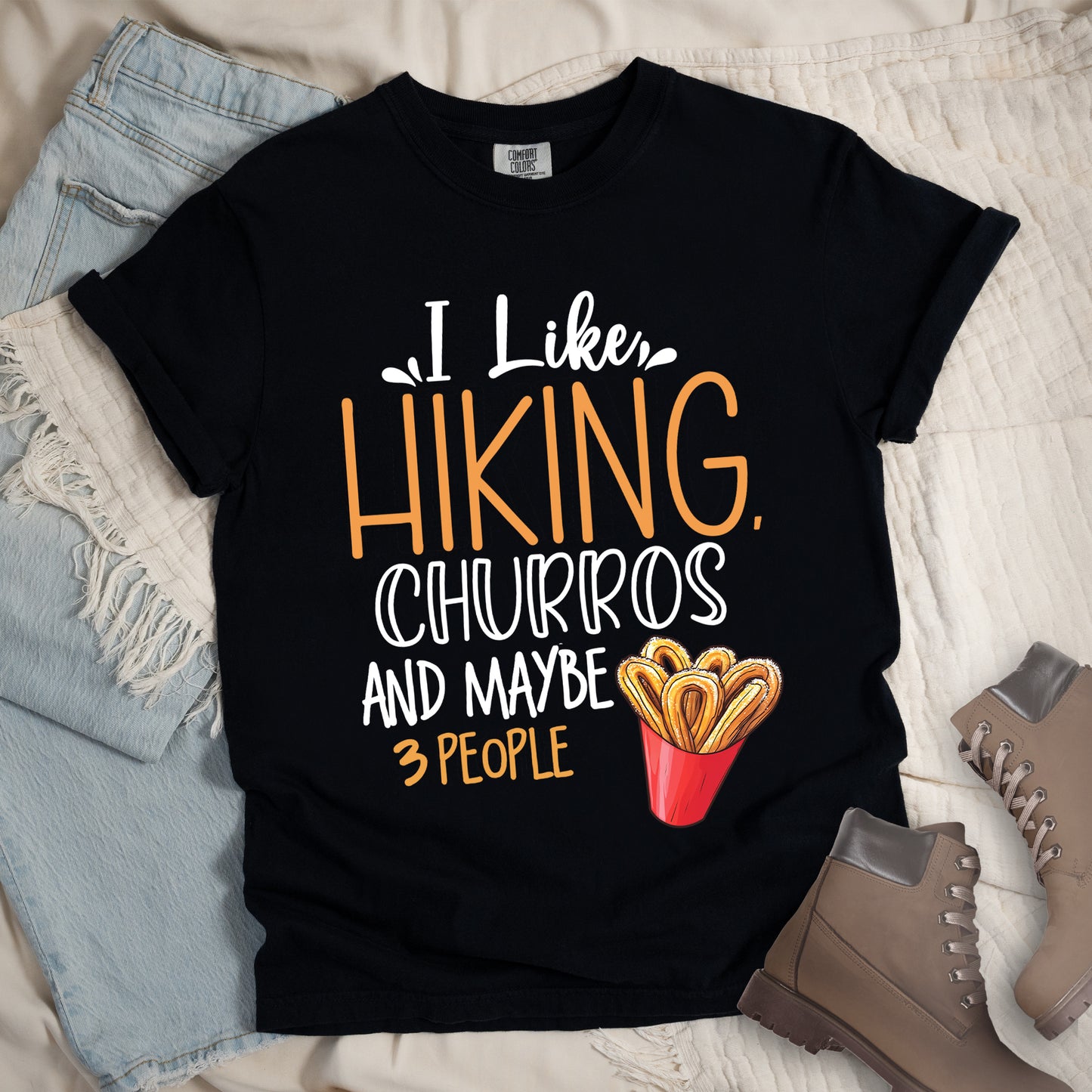 Black t-shirt with white and orange text reading "I Like HIKING, CHURROS AND MAYBE 3 PEOPLE". "I Like" appears in white script at the top, "HIKING," in large orange letters, "CHURROS" in white outlined letters, "AND MAYBE" in white, and "3 PEOPLE" in small orange text at the bottom. Features an illustration of churros in a red container on the right side. Features a classic crew neck and casual rolled sleeves, styled alongside light wash denim and brown hiking boots.