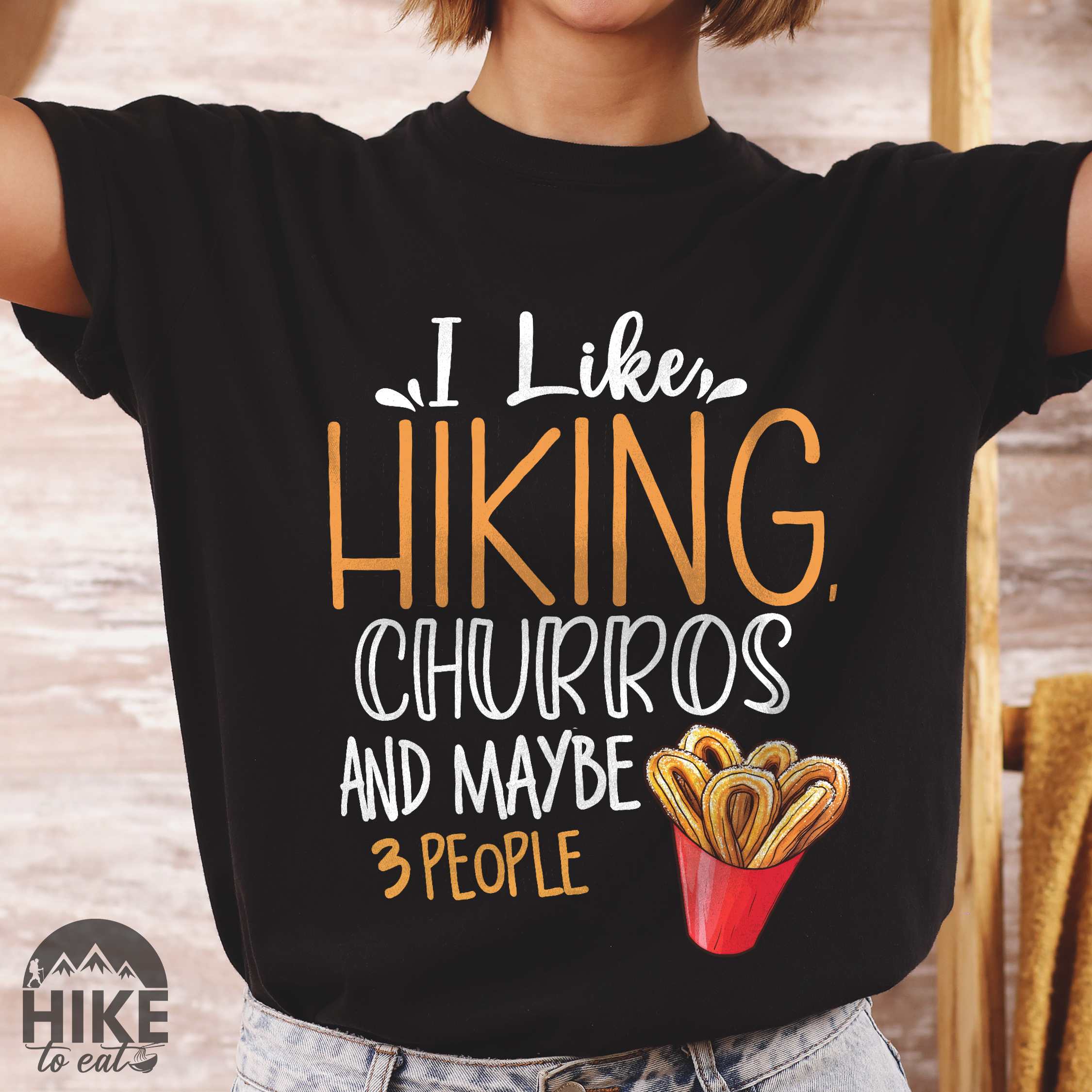 Woman wearing black "I like hiking, churros and maybe three people " colored vintage tee with bold typography, mountains, illustrated a serving of Churros, and backpackers with a sweet tooth.