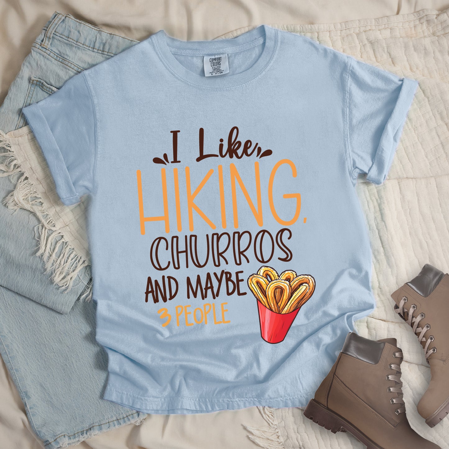 Chambray (Light Blue) t-shirt with deep brown and orange text reading "I Like HIKING, CHURROS AND MAYBE 3 PEOPLE". "I Like" appears in deep brown script at the top, "HIKING," in large orange letters, "CHURROS" in deep brown outlined letters, "AND MAYBE" in deep brown, and "3 PEOPLE" in small orange text at the bottom. Features an illustration of churros in a red container on the right side. Features a classic crew neck and casual rolled sleeves, styled alongside light wash denim and brown hiking boots.