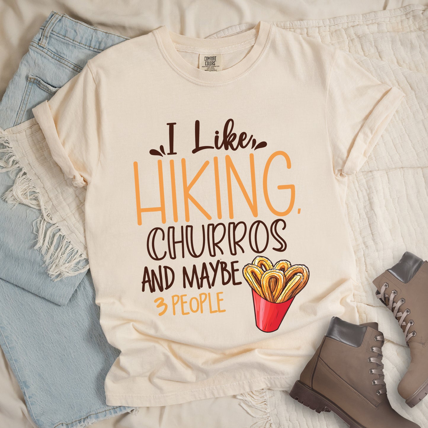 Ivory (Cream White) t-shirt with deep brown and orange text reading "I Like HIKING, CHURROS AND MAYBE 3 PEOPLE". "I Like" appears in deep brown script at the top, "HIKING," in large orange letters, "CHURROS" in deep brown outlined letters, "AND MAYBE" in deep brown, and "3 PEOPLE" in small orange text at the bottom. Features an illustration of churros in a red container on the right side. Features a classic crew neck and casual rolled sleeves, styled alongside light wash denim and brown hiking boots.